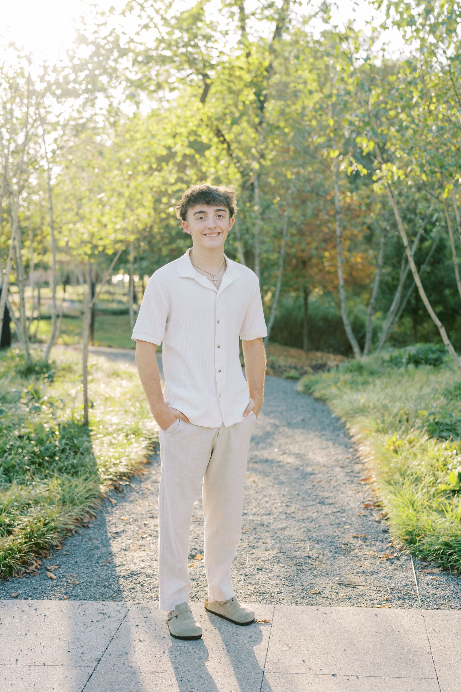 A person in light colored clothing stands on a sunlit gravel path surrounded by green trees and foliage.
