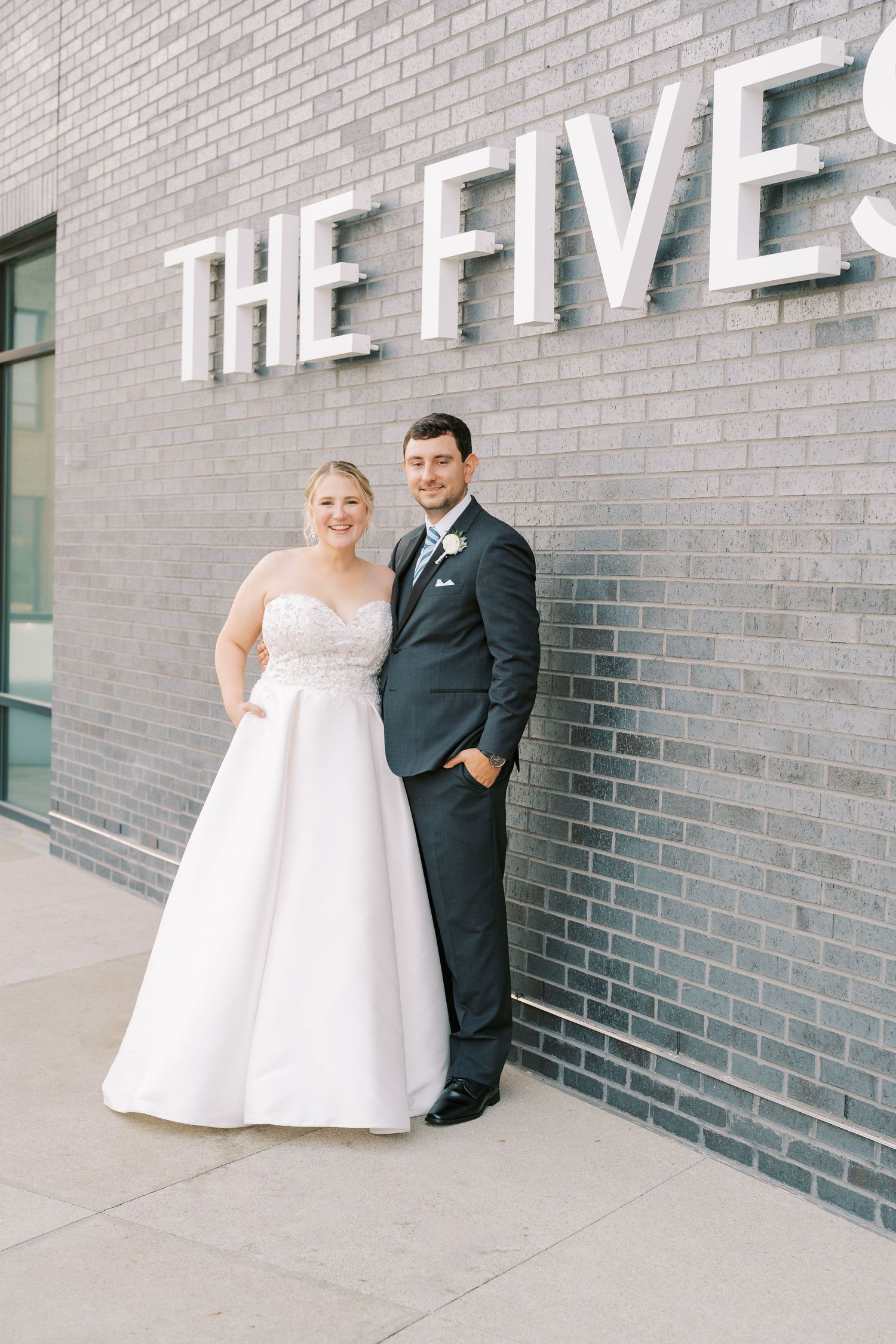 A couple in wedding attire poses against a modern gray brick wall in a series of elegant portraits.