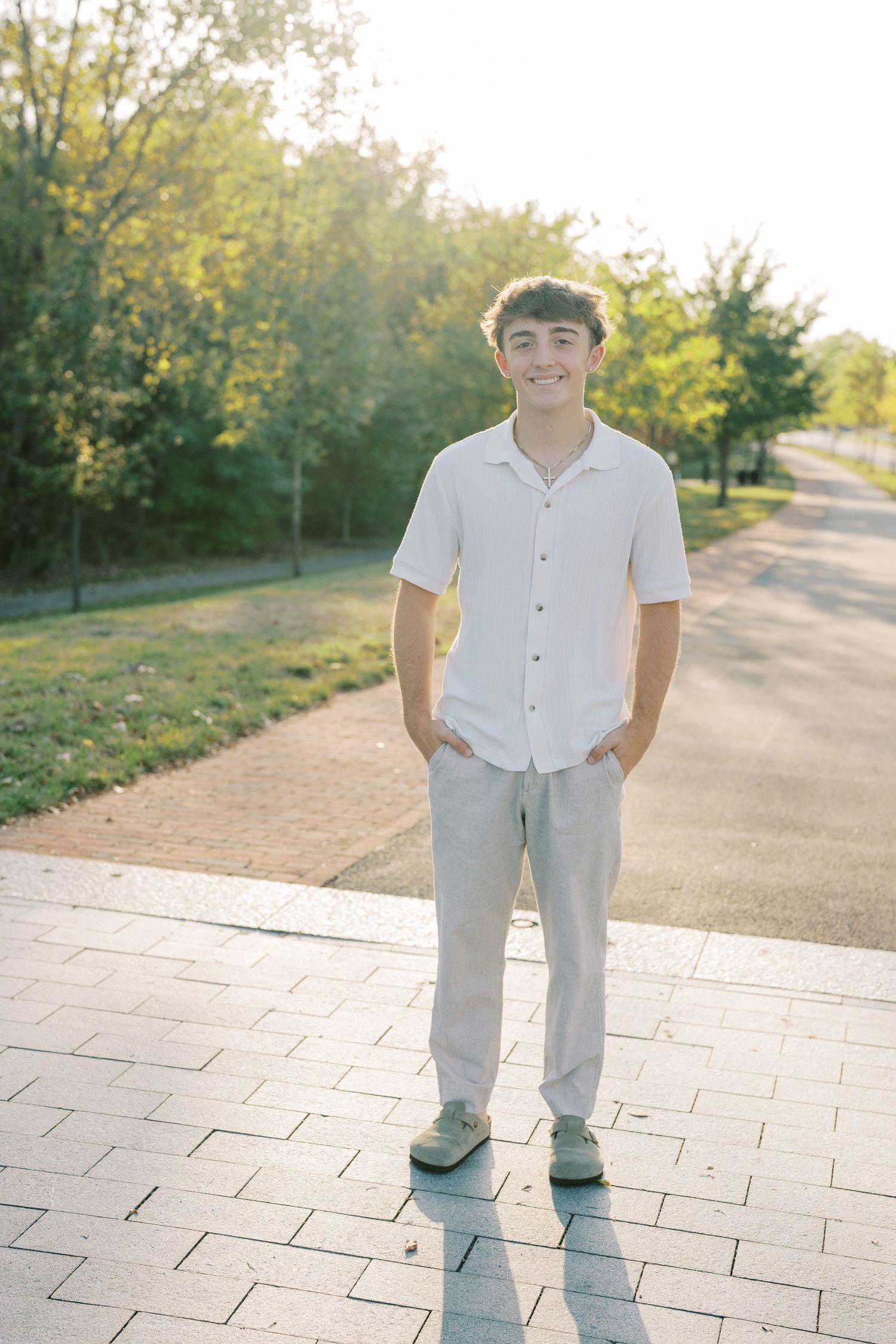 A person wearing a white button-down shirt and gray pants stands on a stone pathway in a sunlit outdoor setting.