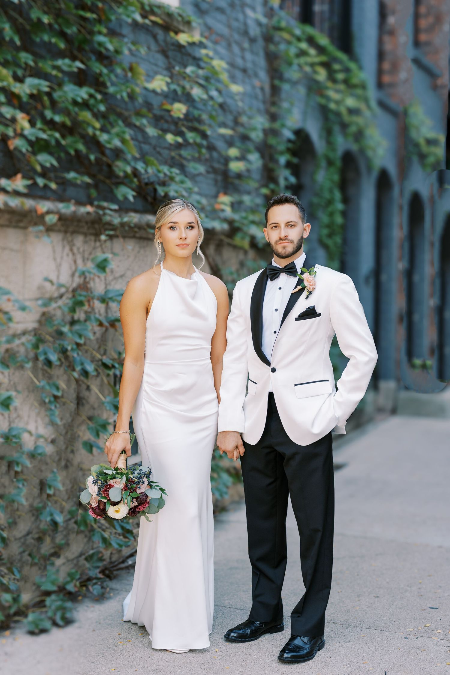 A romantic wedding photo series showing a couple in white and black formal attire posing in an ivy-covered alley.