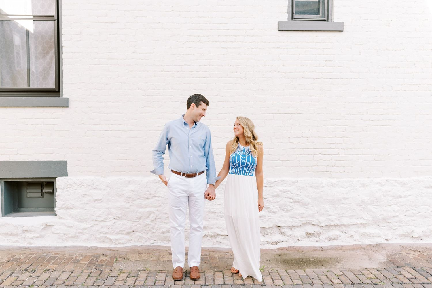 A couple in white outfits walks together on a brick sidewalk beside a white building on a sunny day.