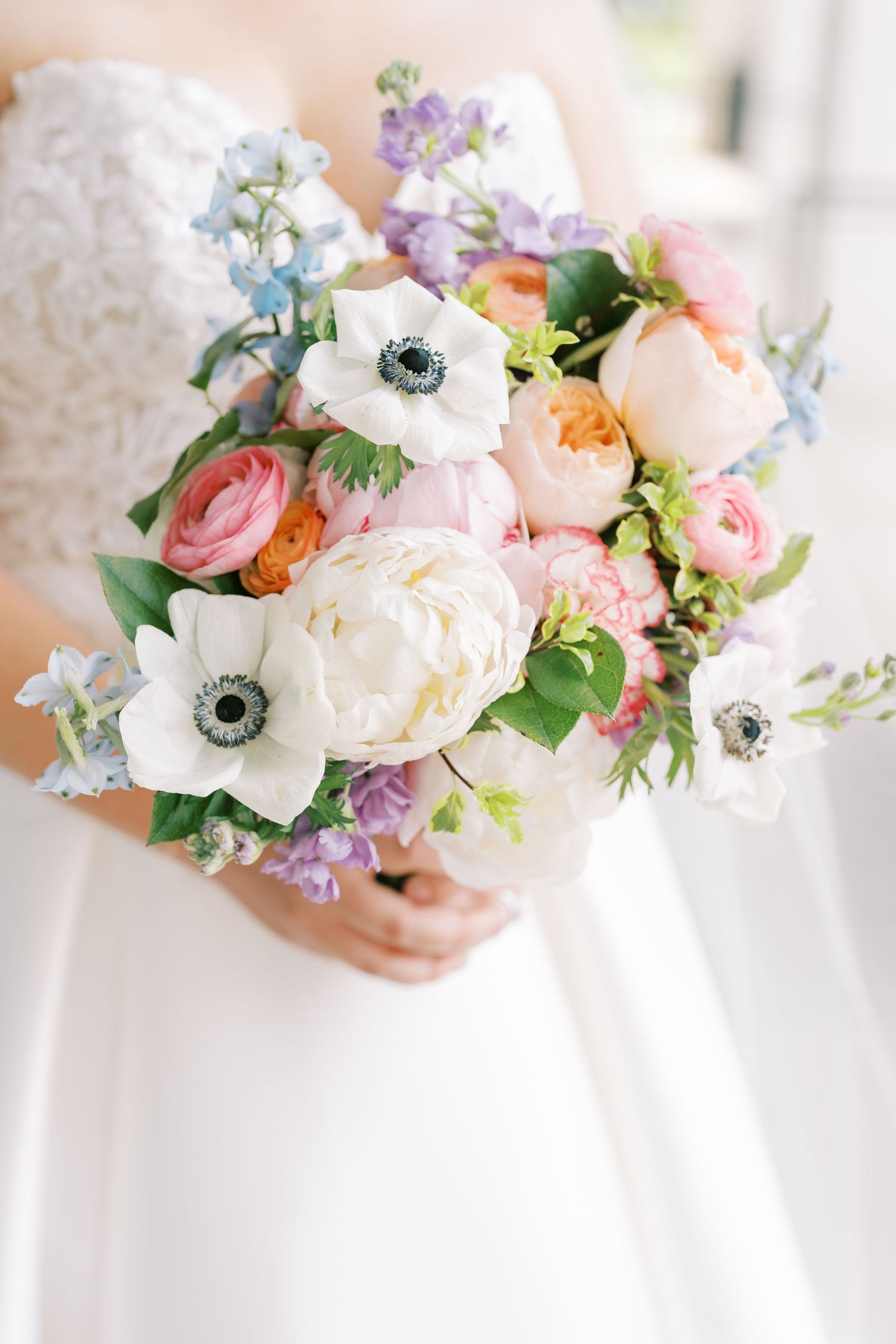 A delicate spring wedding bouquet featuring white anemones, peach garden roses, pink ranunculus and blue delphinium flowers.