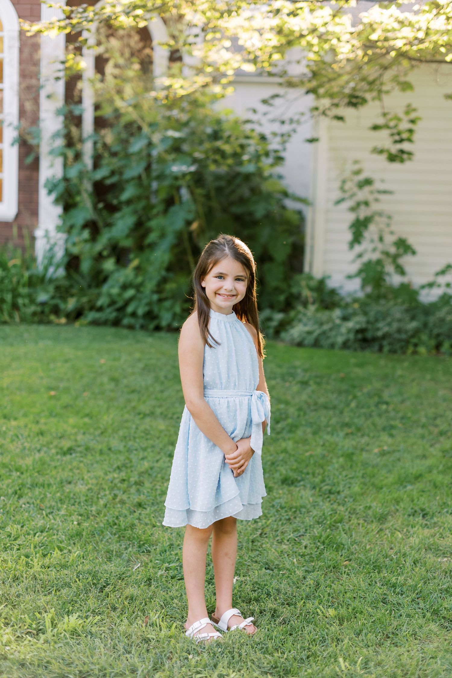 Child in light blue dress standing on grass near garden.