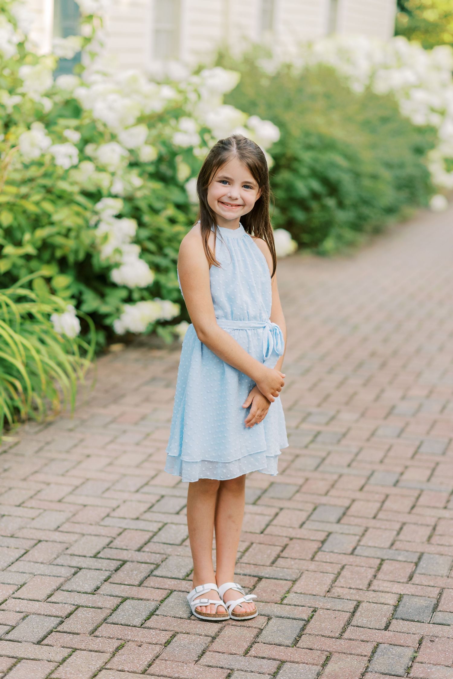 Child in light blue summer dress standing on brick pathway with white flowers in background.