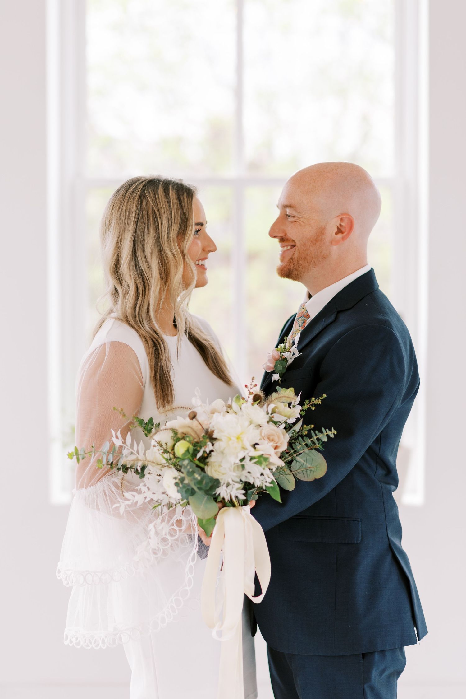 Wedding couple sharing joyful moments in a bright white room with floral bouquet.