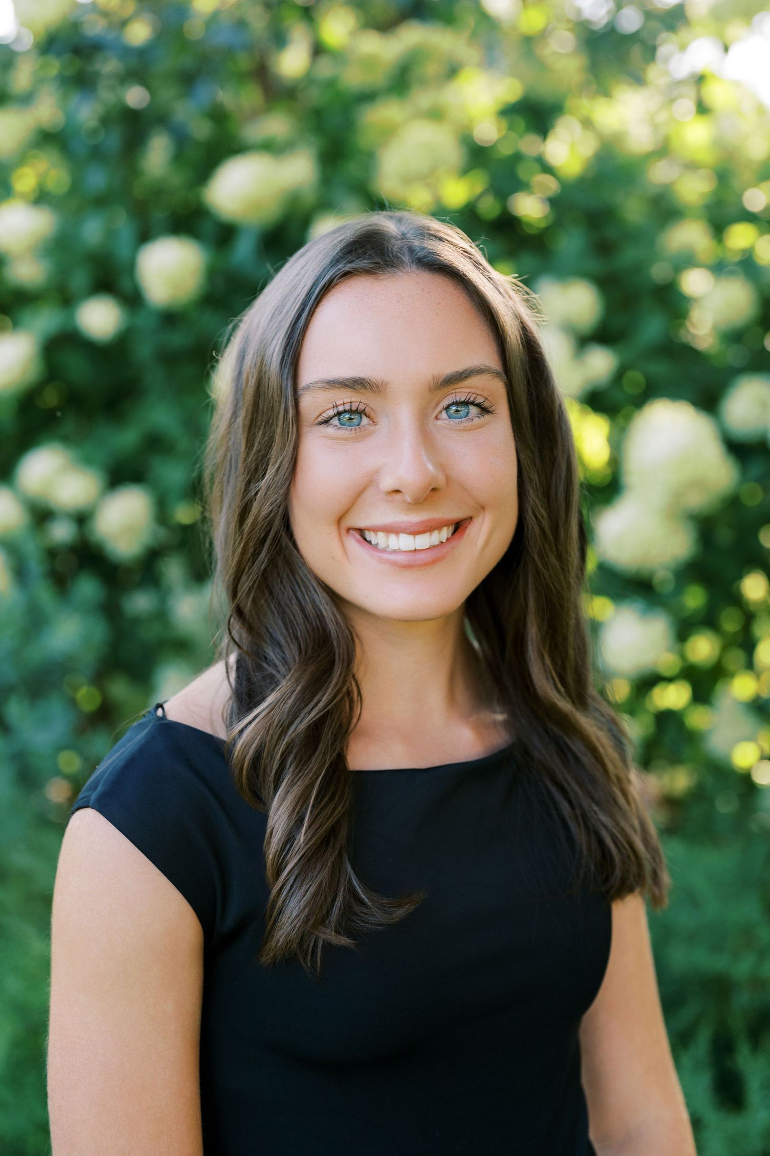 Professional portrait of someone in a black top smiling outdoors with greenery and white flowers in the background.