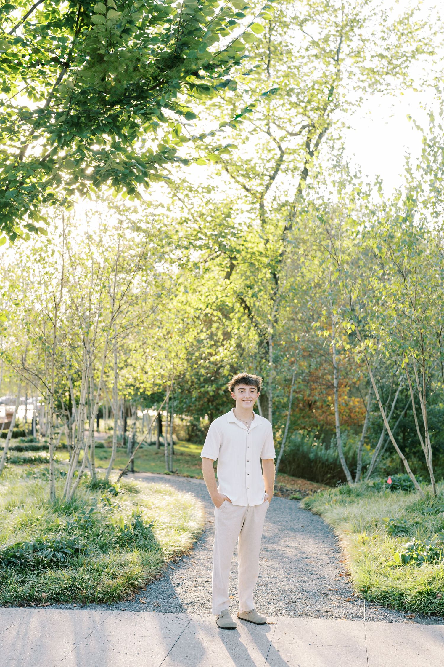 A couple walks along a sunlit garden path surrounded by lush green trees and foliage on a bright summer day.