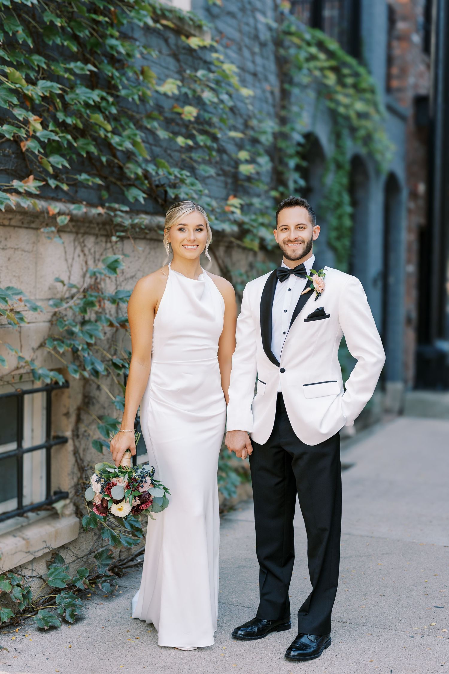 A couple in elegant white and black wedding attire pose together in an outdoor urban setting with ivy-covered walls.
