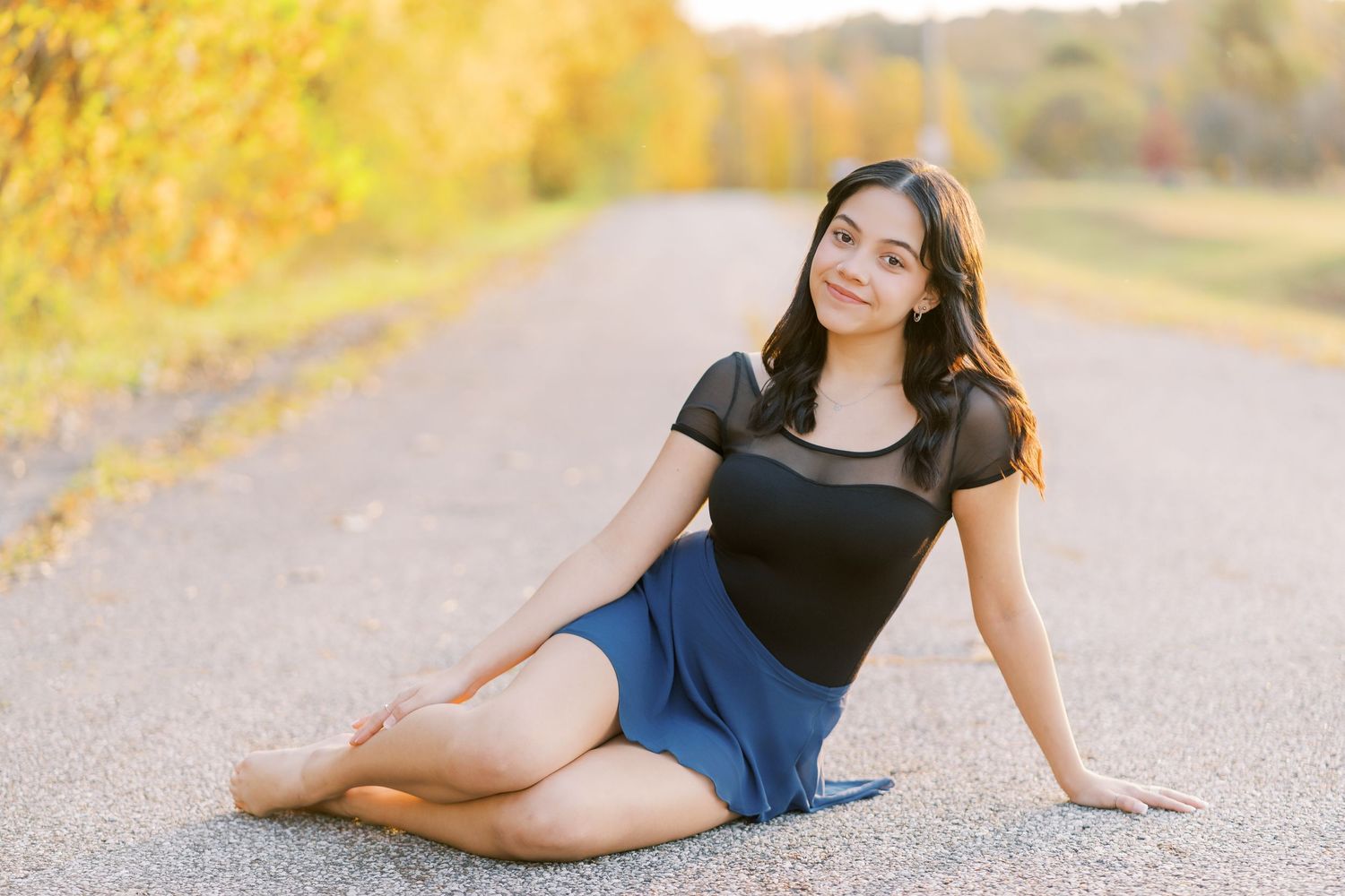 A person in casual attire poses outdoors on a path surrounded by golden autumn foliage during a sunny afternoon.