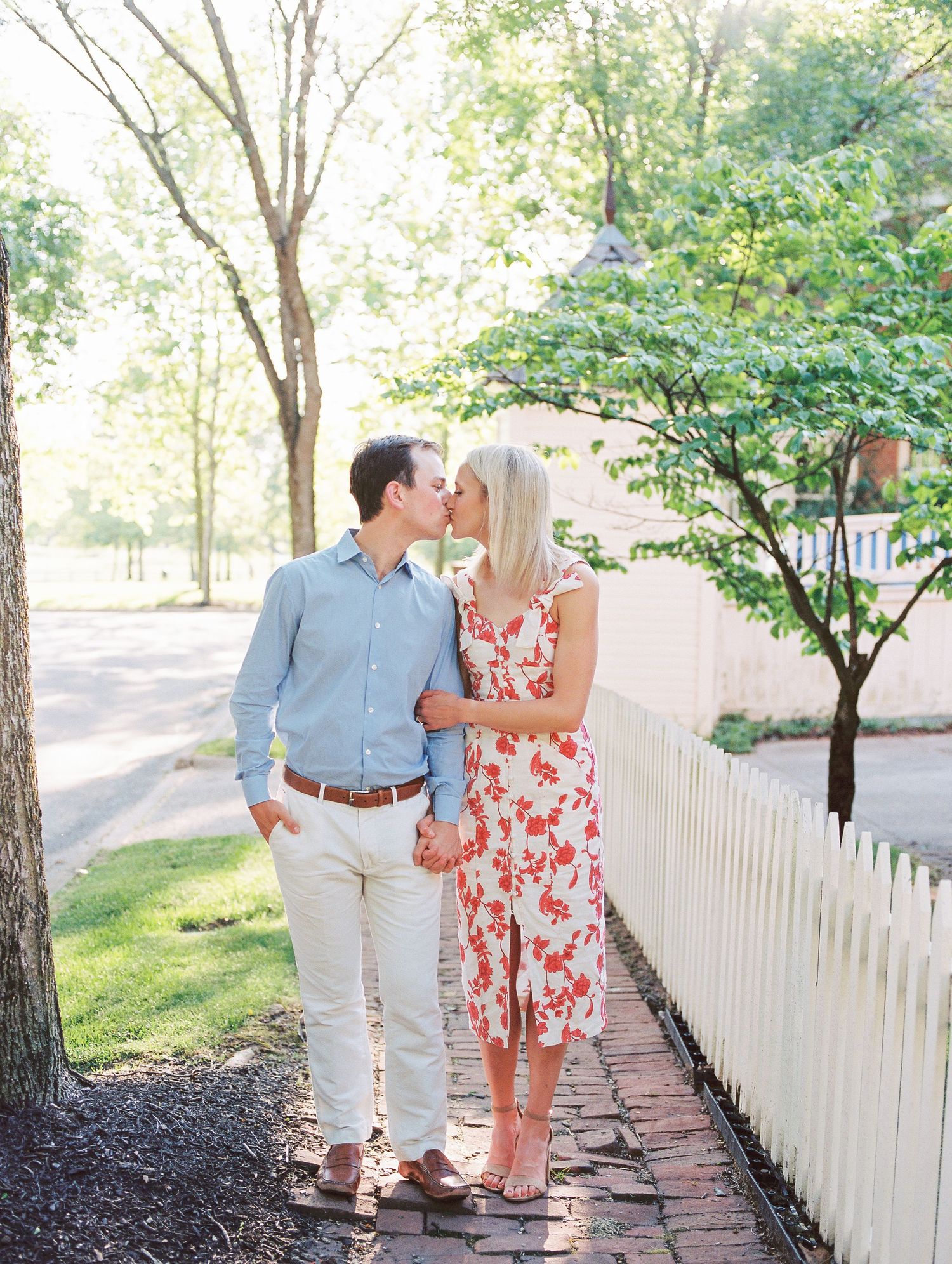 A couple shares a romantic moment on a brick pathway beside a white picket fence on a sunny spring day.