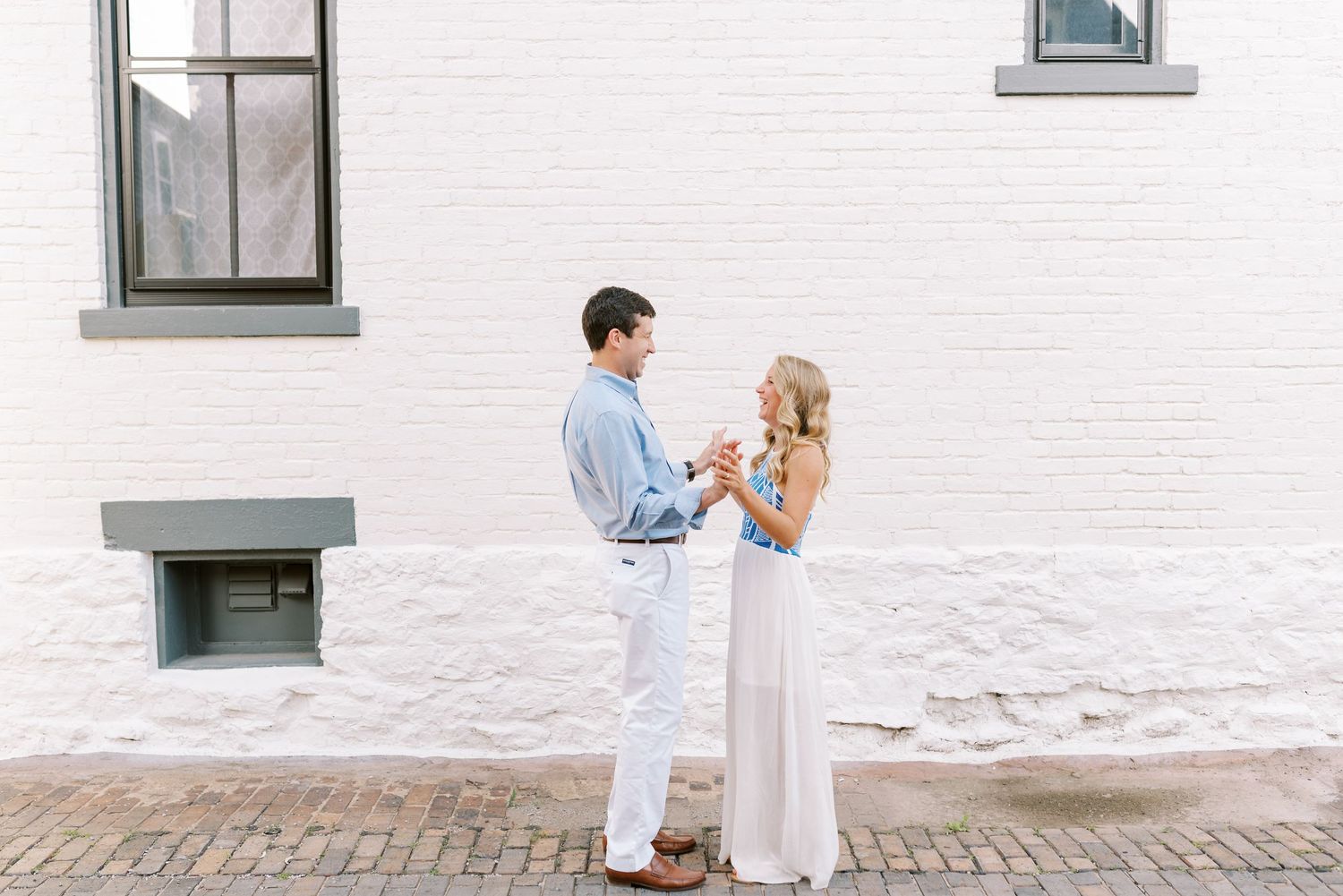 A couple in white outfits poses together against a white building wall with green window frames.