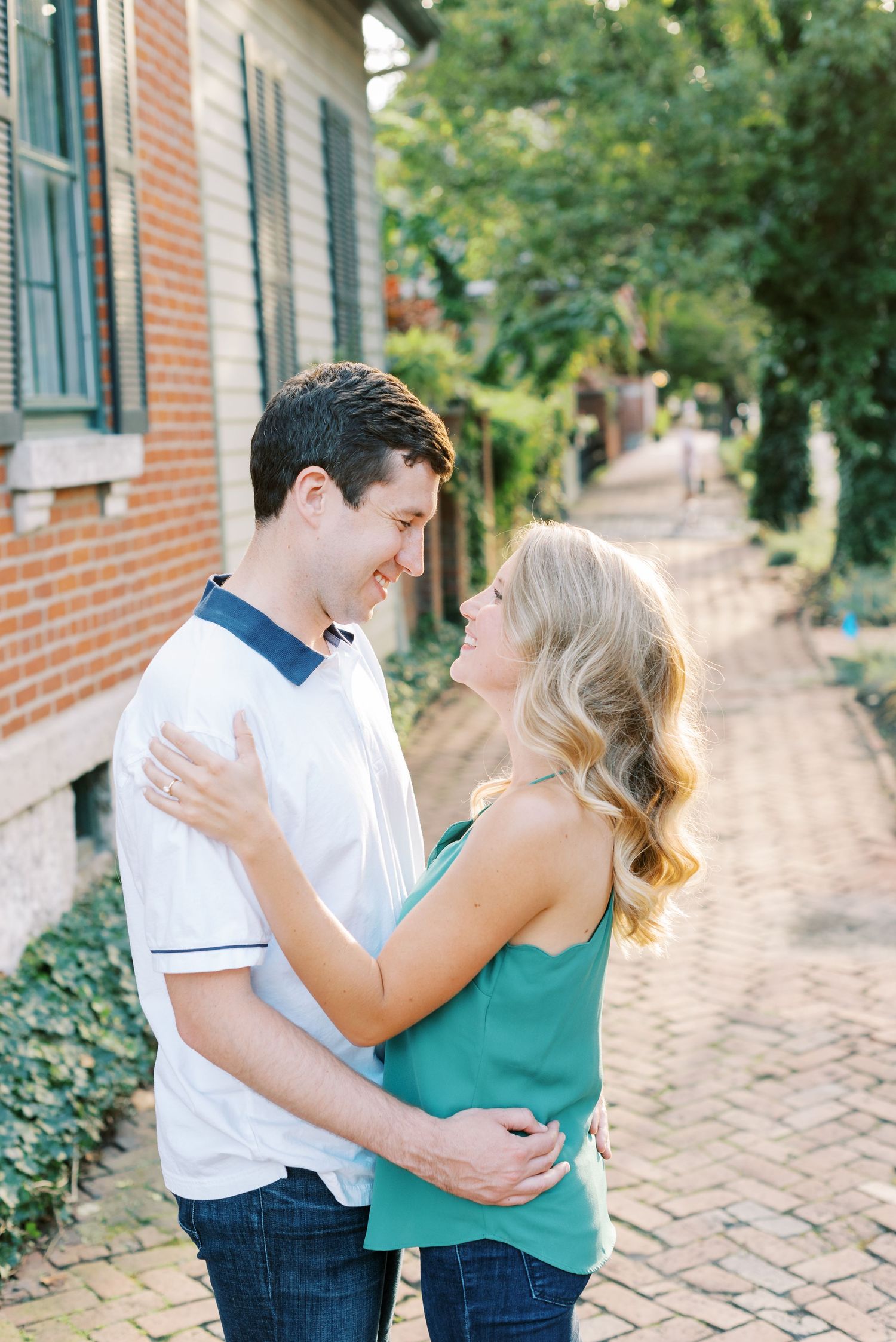 A couple shares an intimate moment on a brick sidewalk next to a historic building with green foliage in the background.