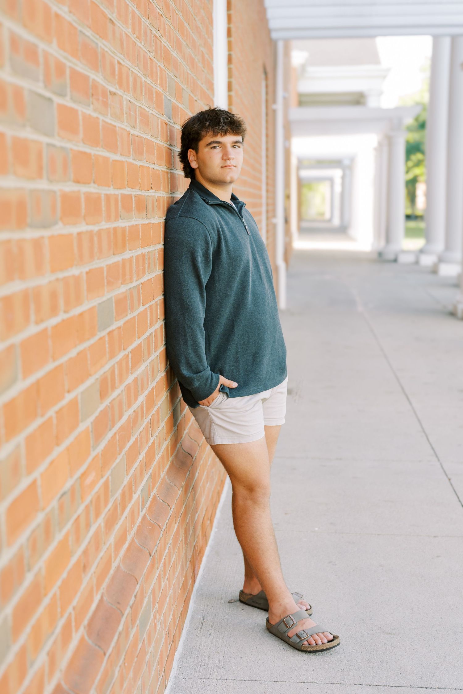 A person in a teal shirt leans against an orange brick wall in a covered walkway with natural lighting.