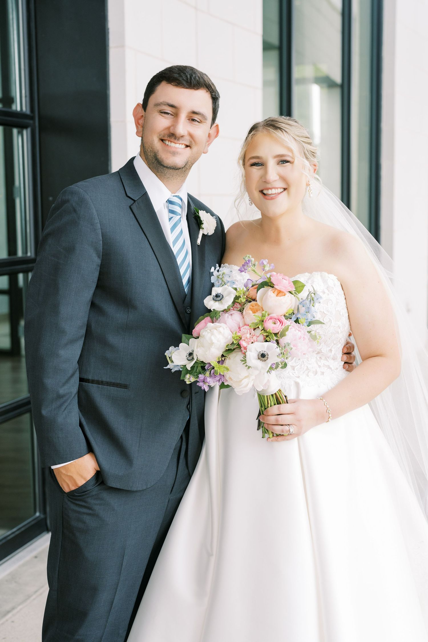A bride and groom pose together on their wedding day, with the bride holding a beautiful pink and white floral bouquet.