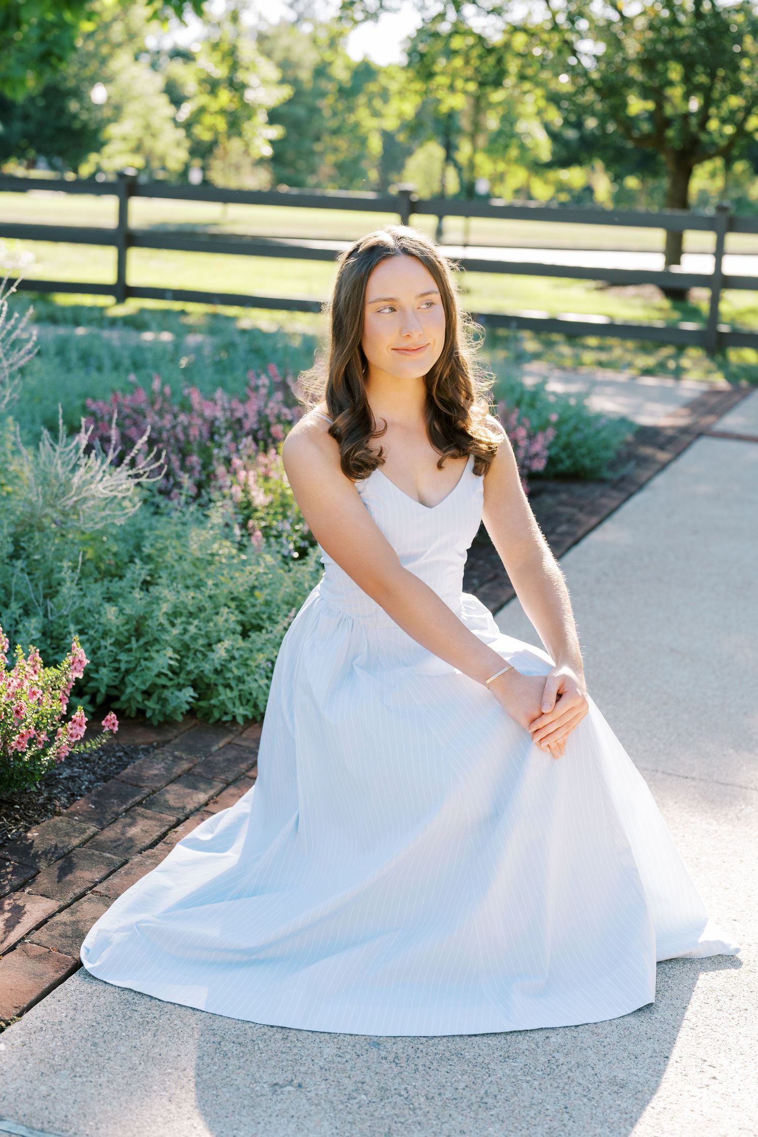 A white wedding gown with lace details photographed in a sunlit garden setting with wooden fencing and blooming flowers.