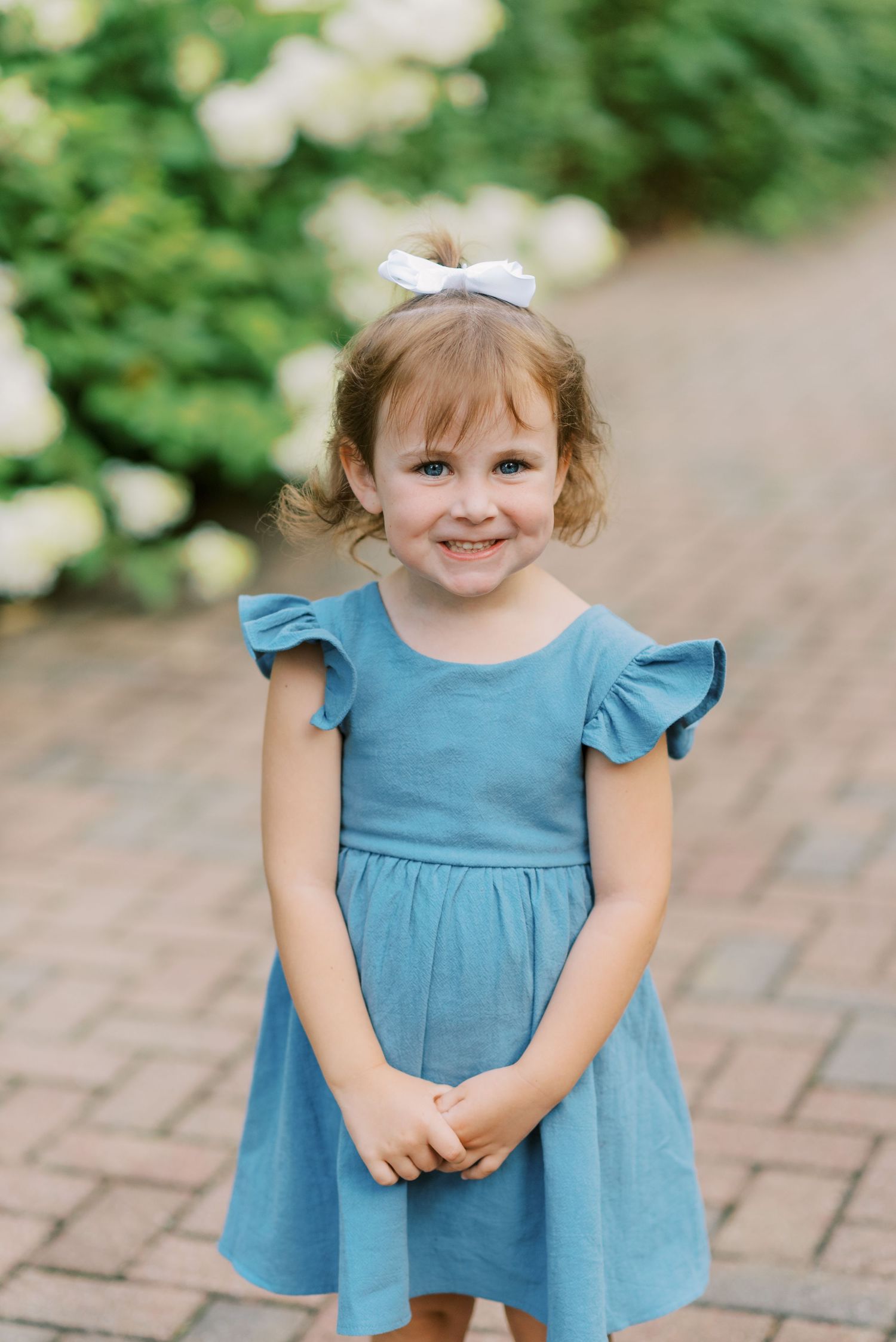 Child wearing blue flutter sleeve dress with white bow accessory standing on brick pathway.