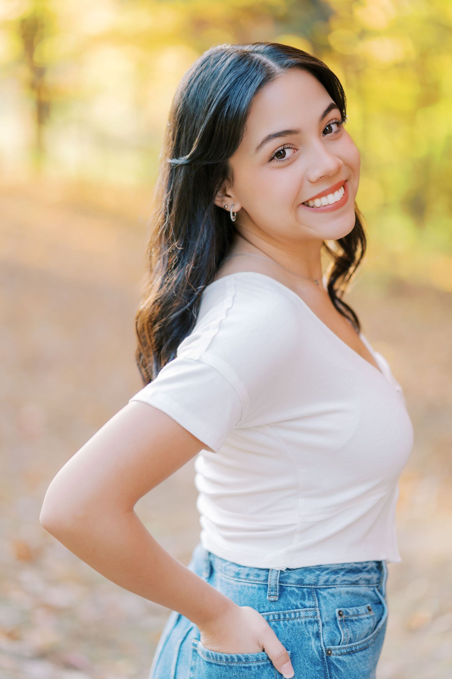 Portrait of someone in a white t-shirt and blue jeans posing outdoors with a bright smile during autumn.
