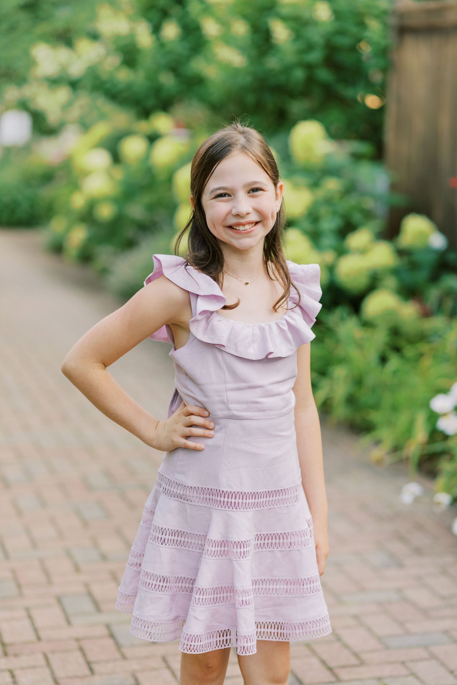 Child in pink eyelet dress posing on brick pathway near garden.