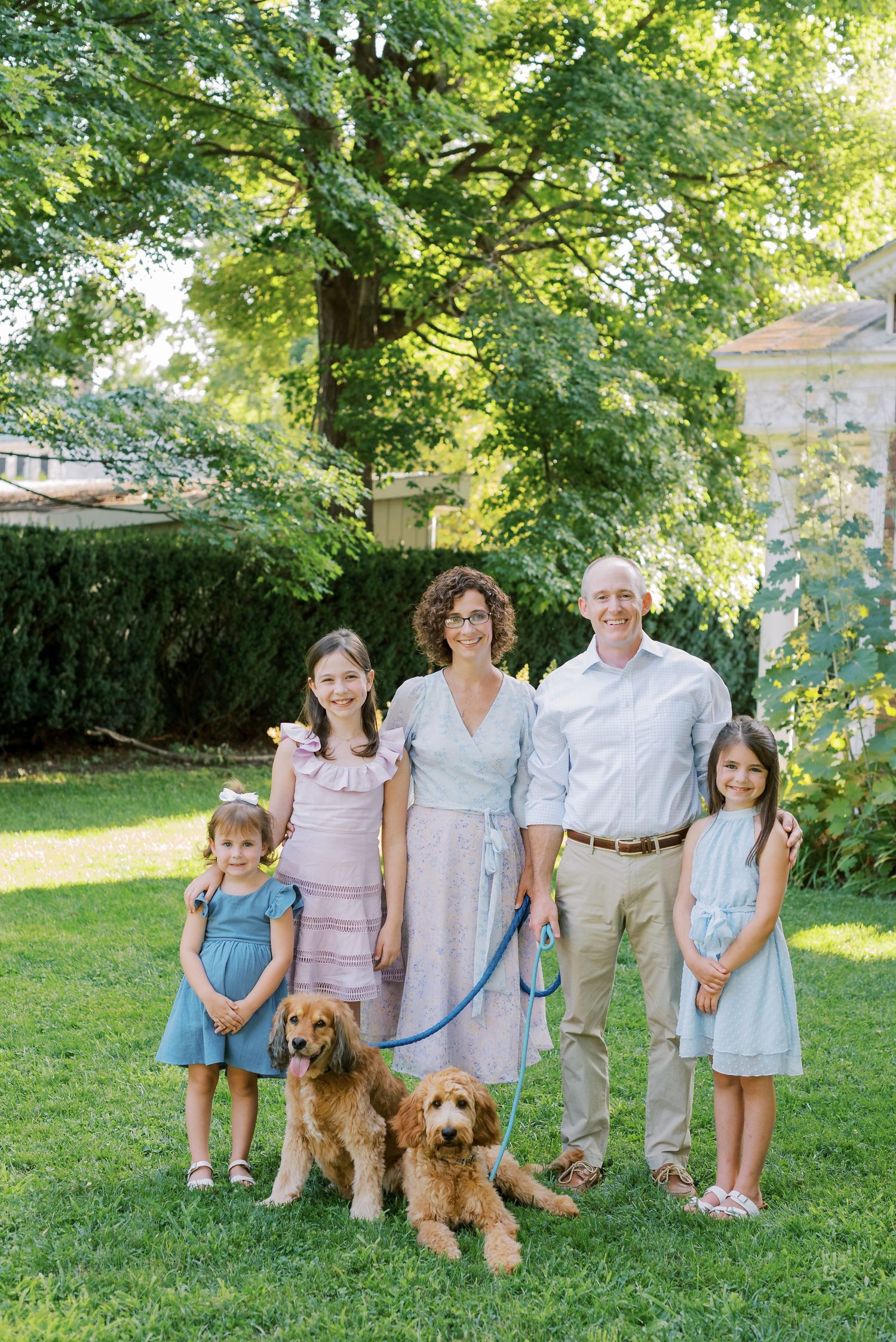 Family members and their pets pose together on a grassy lawn with trees and shrubs in the background.