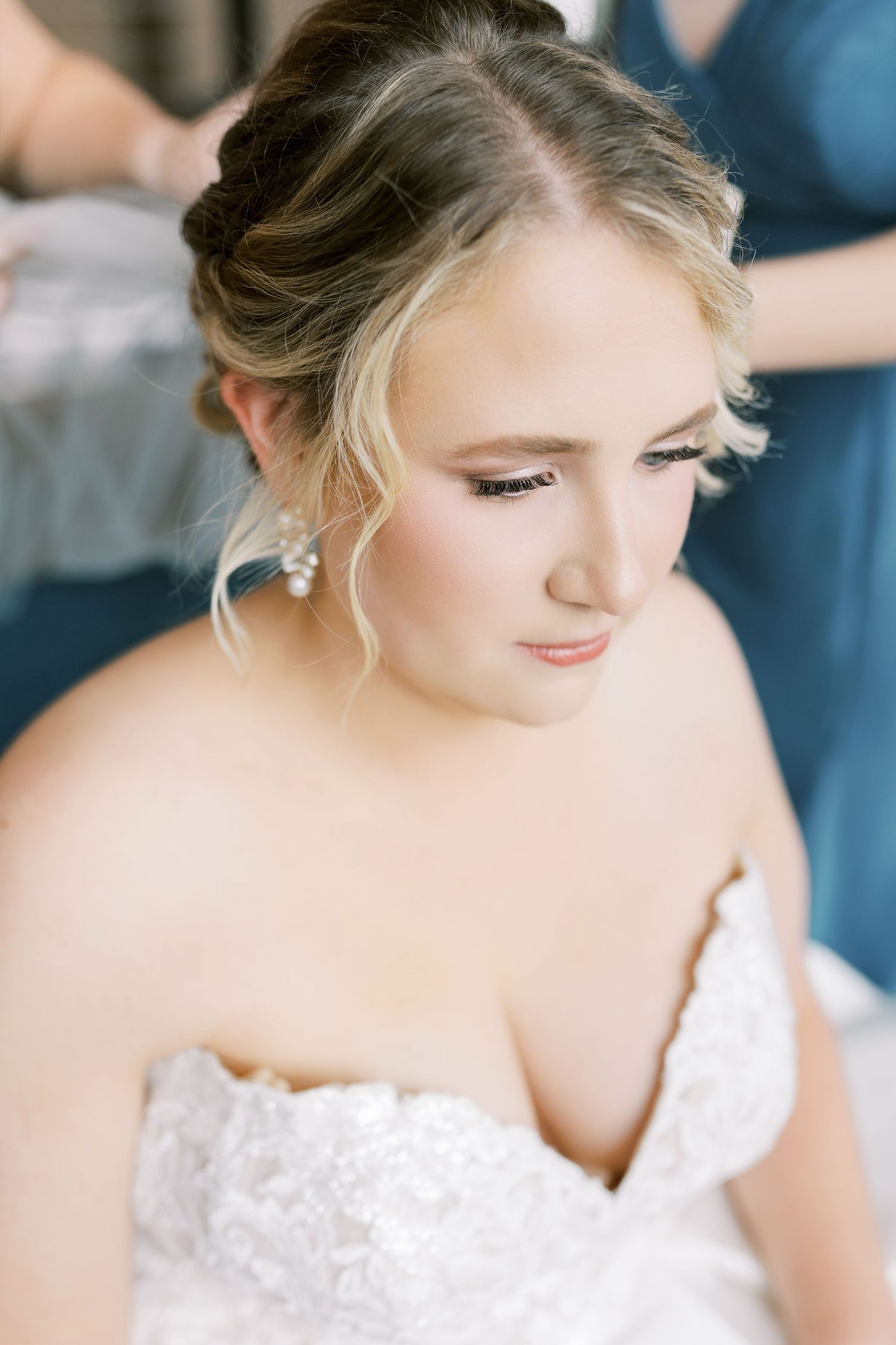 A bride in a white lace wedding dress looks down elegantly while wearing delicate earrings and an upswept hairstyle.