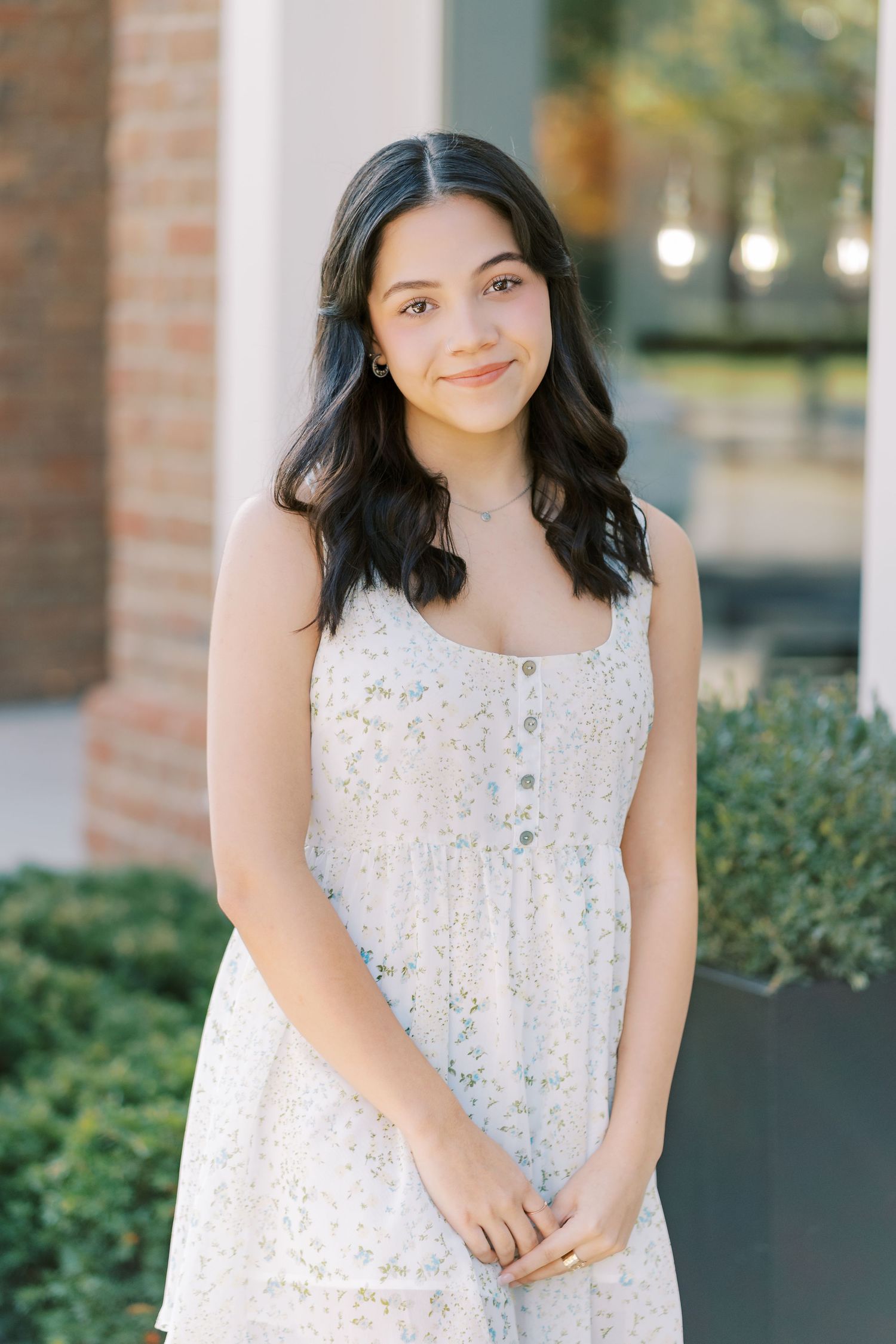 A person in a white floral sleeveless dress smiles warmly while standing against a brick wall outdoors.