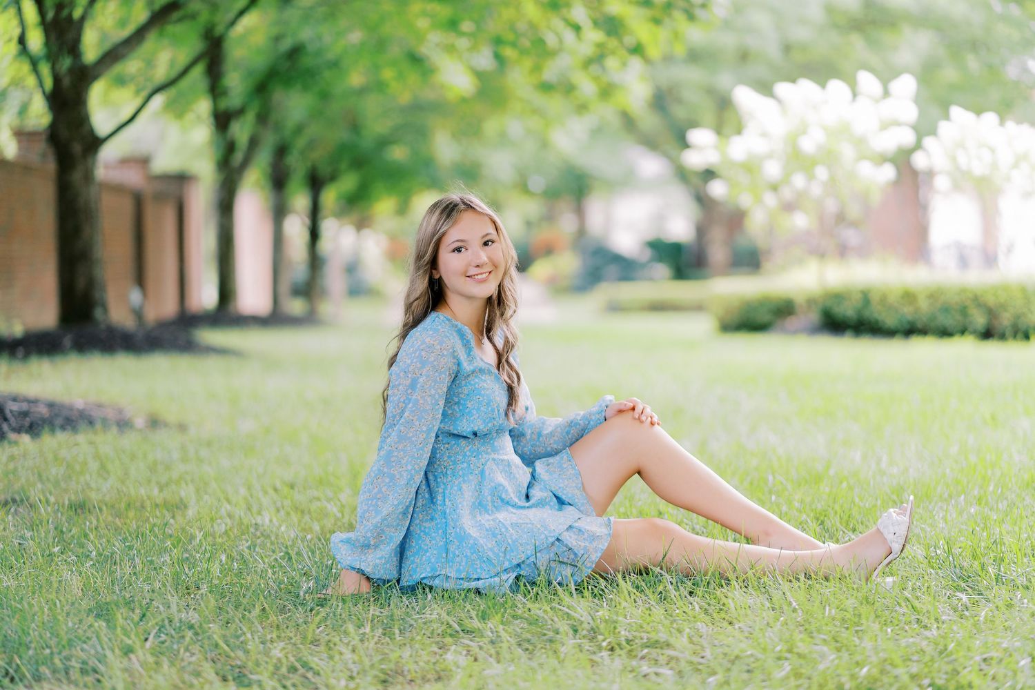 A person in a light blue dress sits on green grass under a row of trees on a sunny spring day.