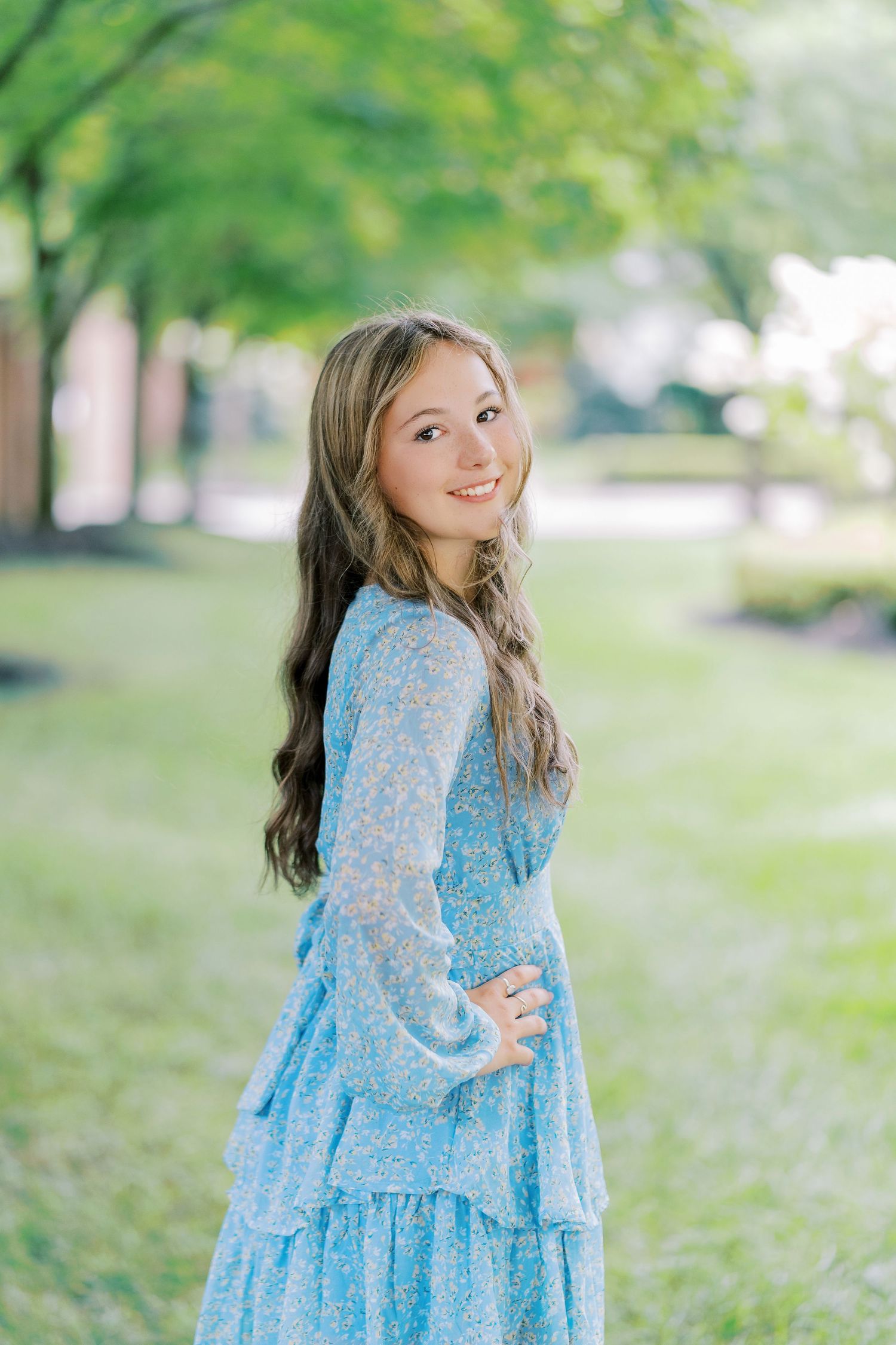 A young girl in a flowing blue lace dress poses gracefully in a sunlit park setting.