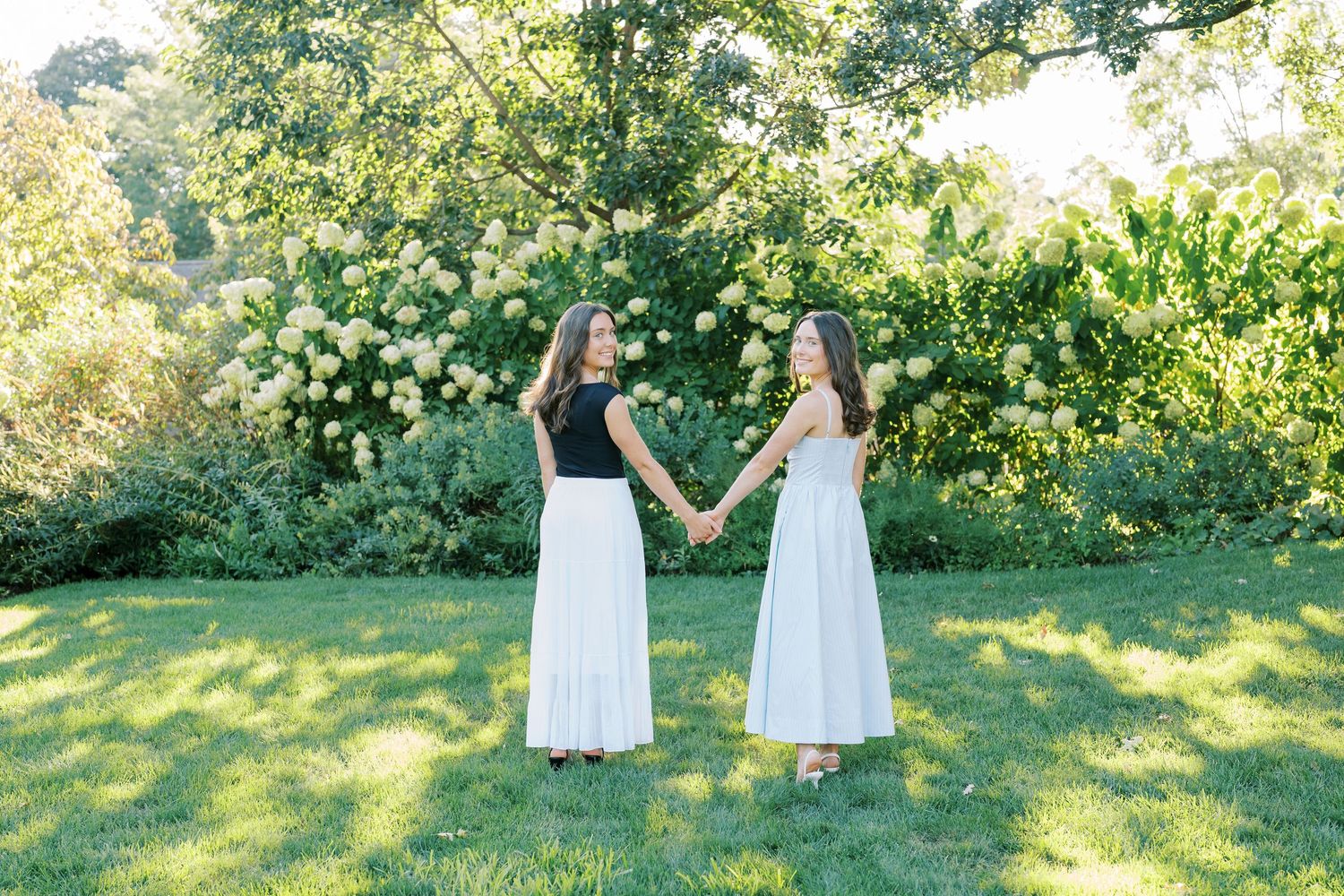 Two people in white dresses dance and twirl together in a sunlit garden setting with lush green foliage.