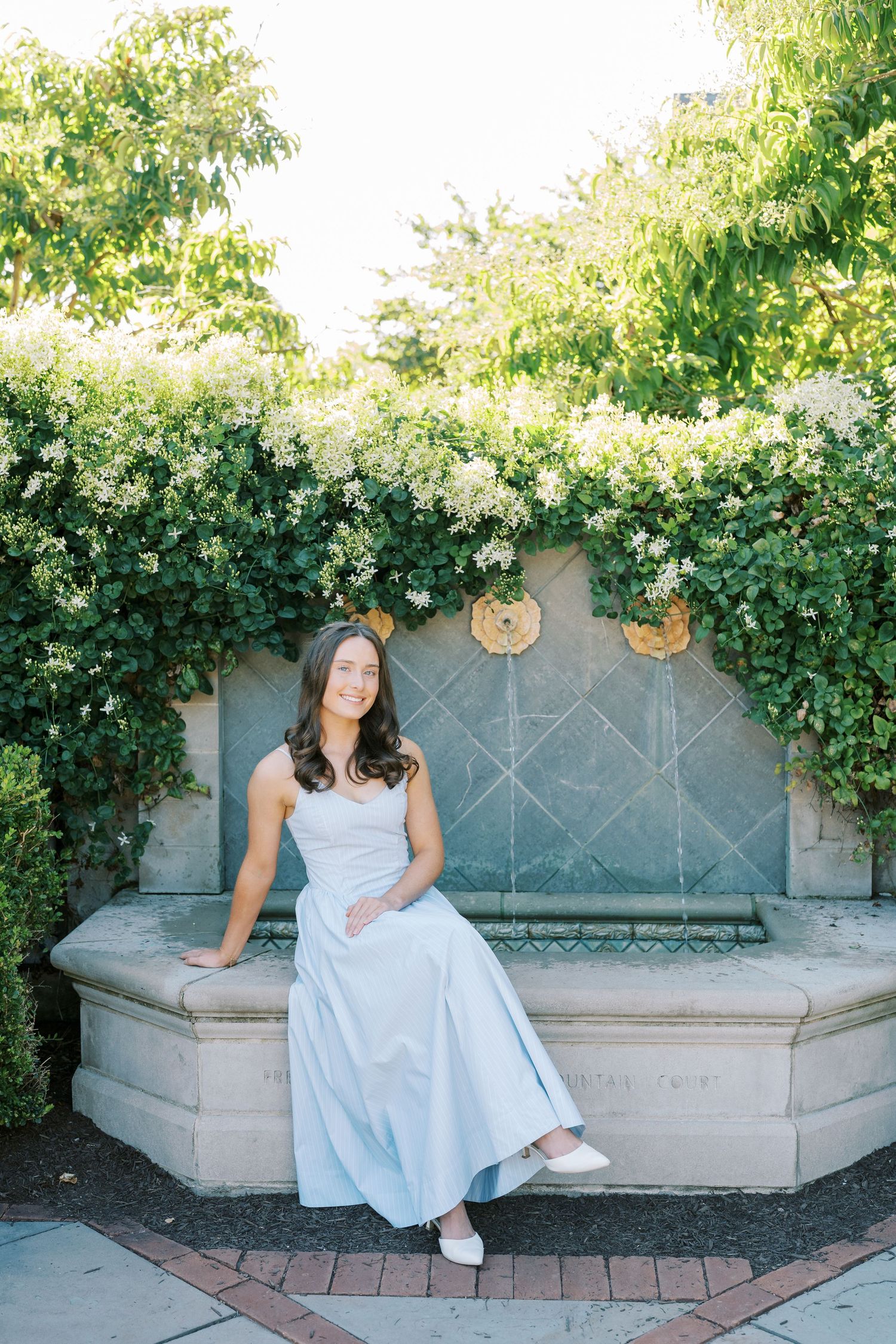 A person in a light blue dress sits on a stone bench surrounded by lush greenery and climbing vines.