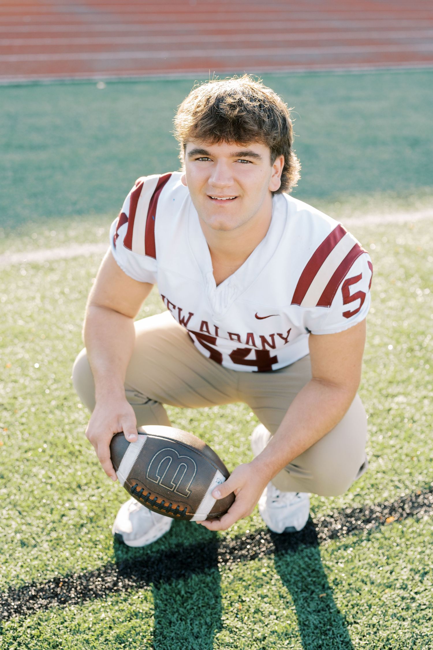Football player in white and maroon uniform crouches on sunny field with ball during practice session.