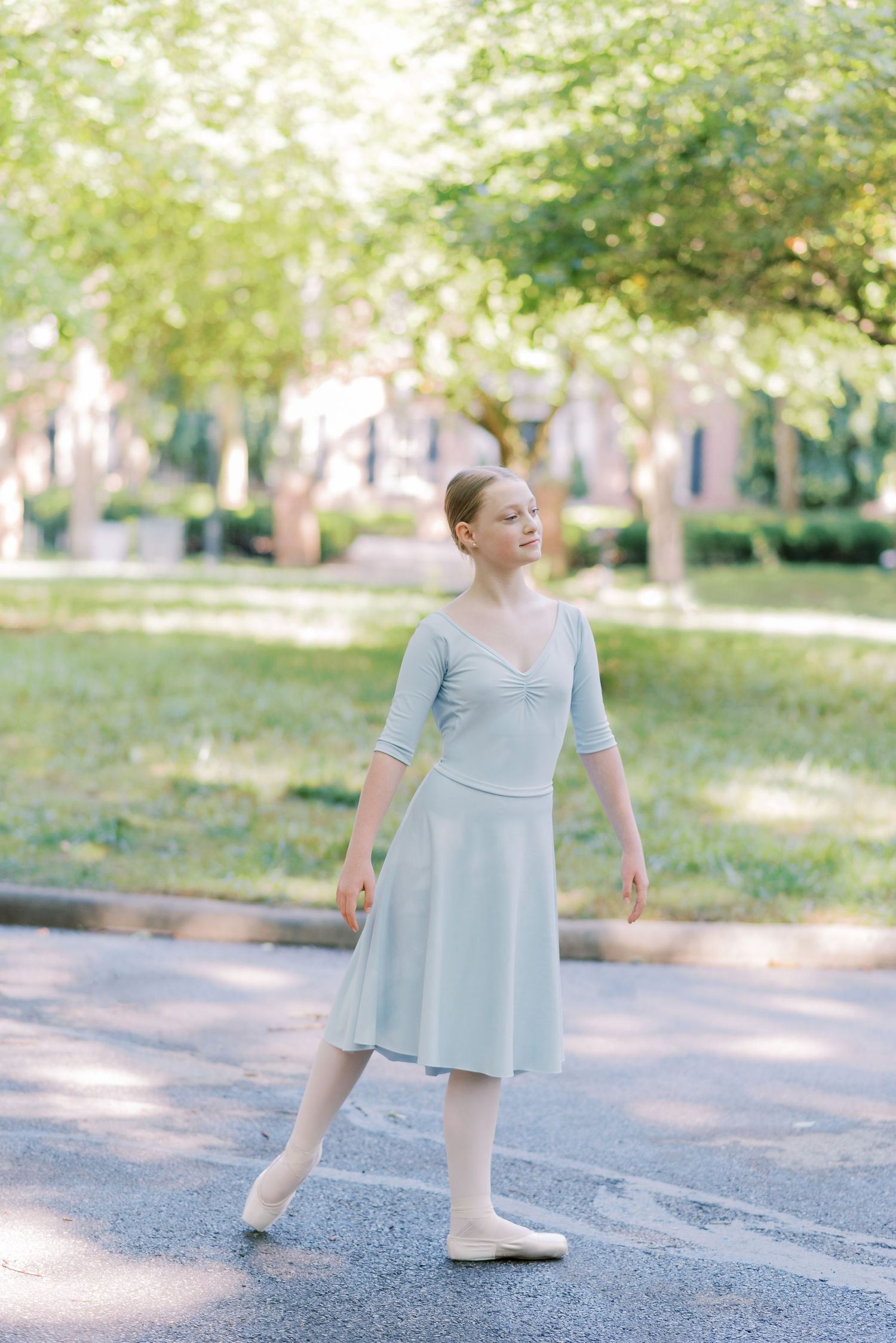 A dancer in a light blue dress performs graceful ballet movements on an outdoor path surrounded by trees.