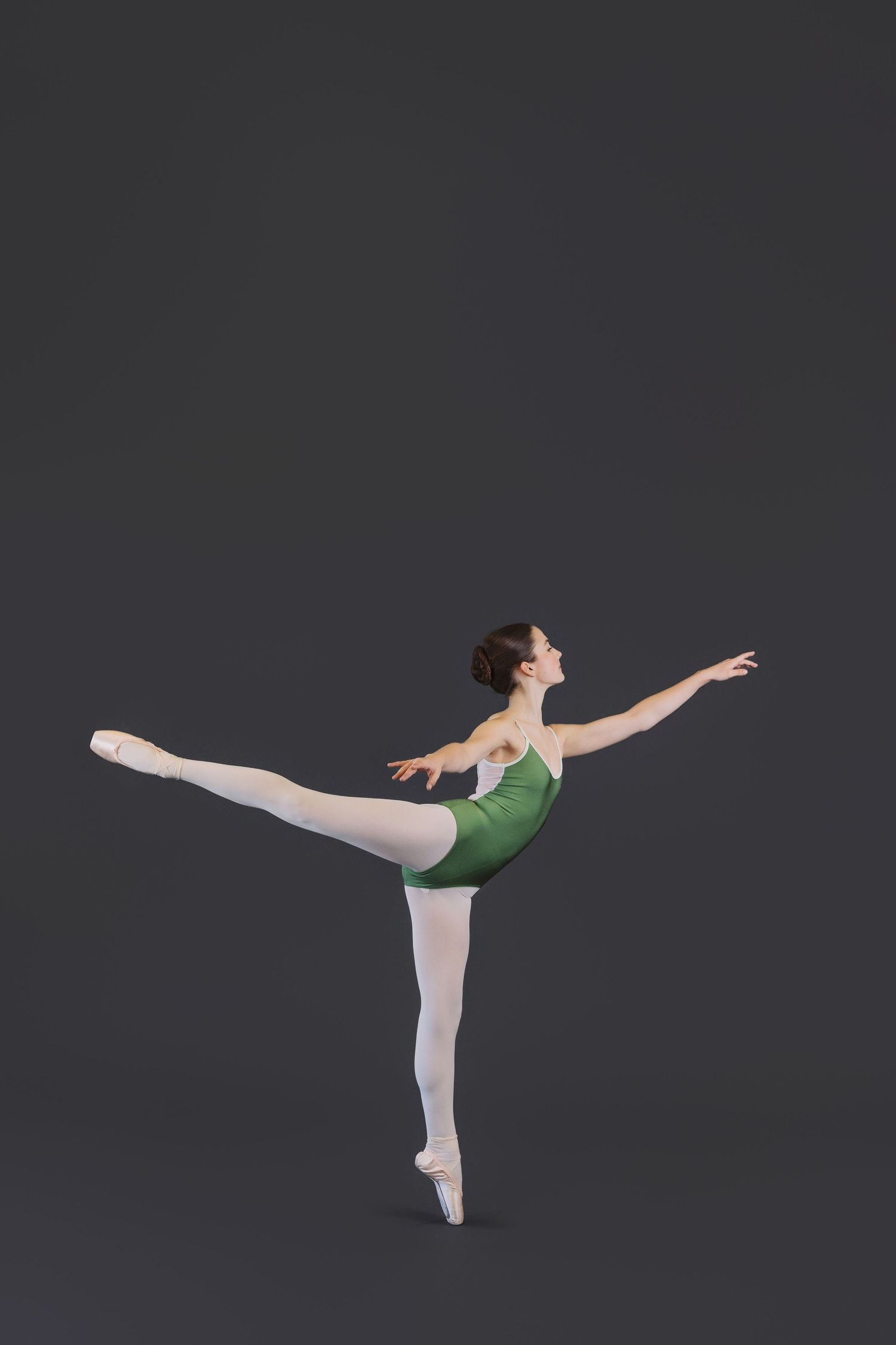 Ballet dancer in green leotard and white tights performs graceful arabesque sequence against dark background.