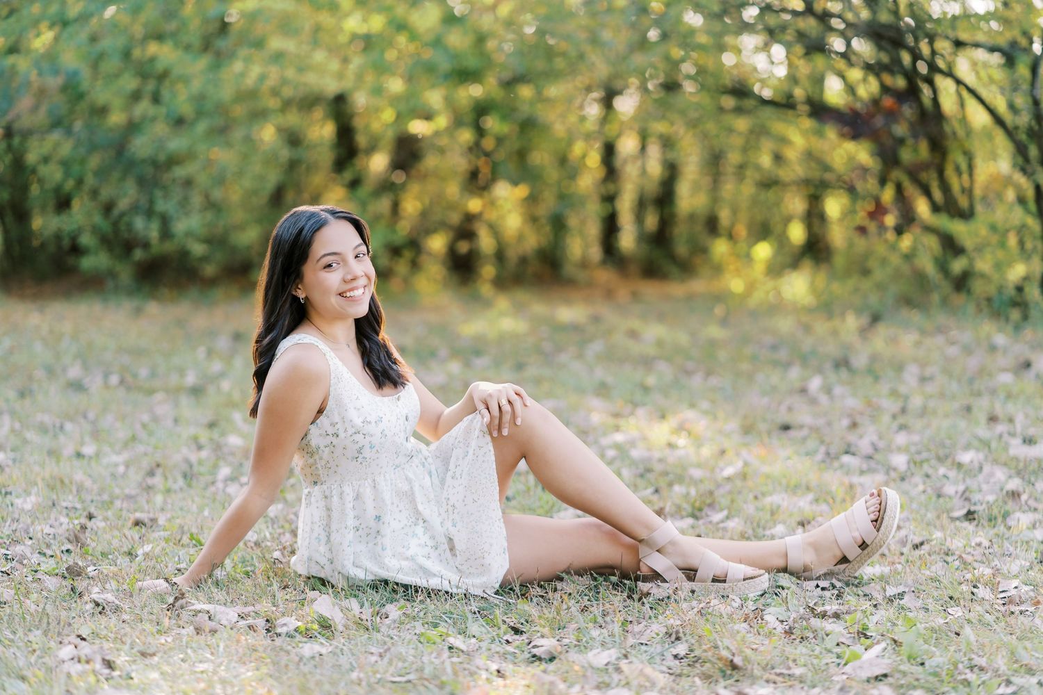A person in a white dress sits gracefully on autumn leaves during an outdoor photoshoot in a sunlit forest.