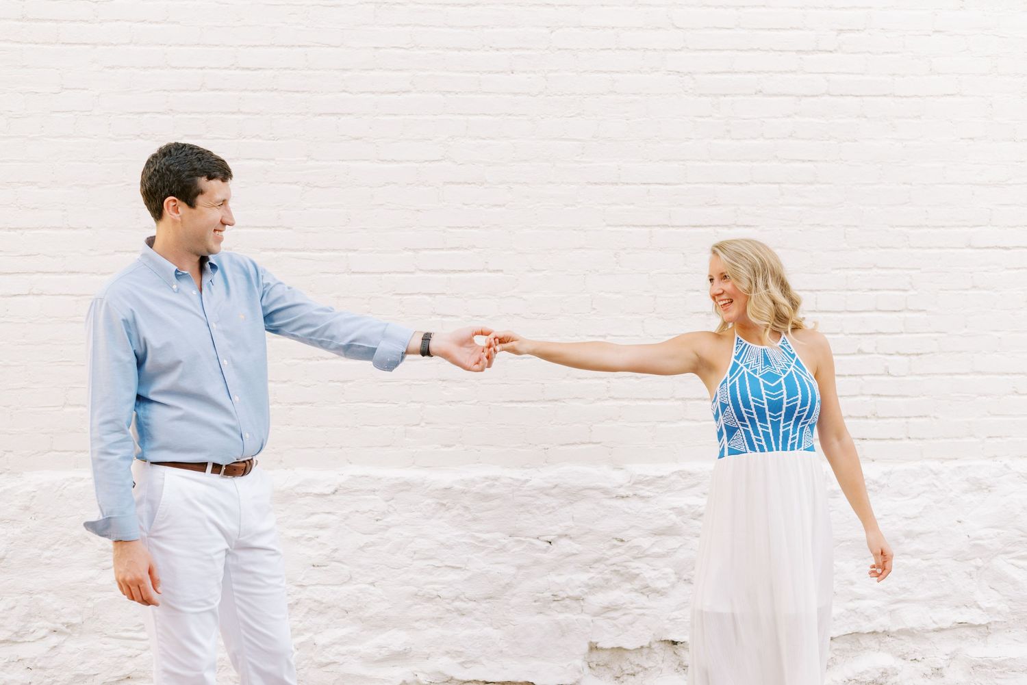A couple in blue and white outfits hold hands while standing against a white wall.
