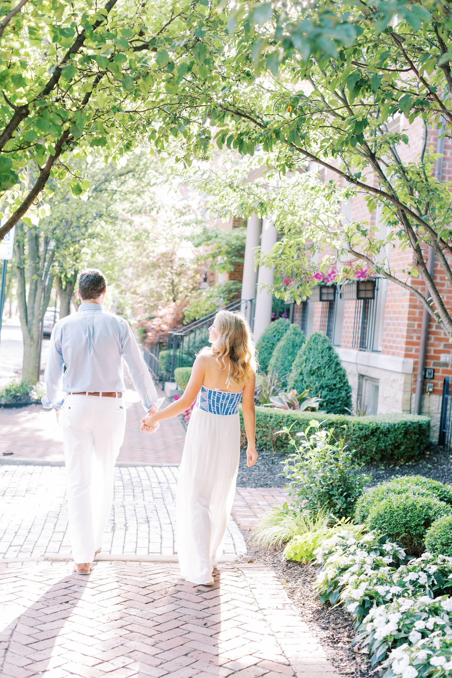 A couple walks down a brick path surrounded by lush green trees and bushes in a garden setting.