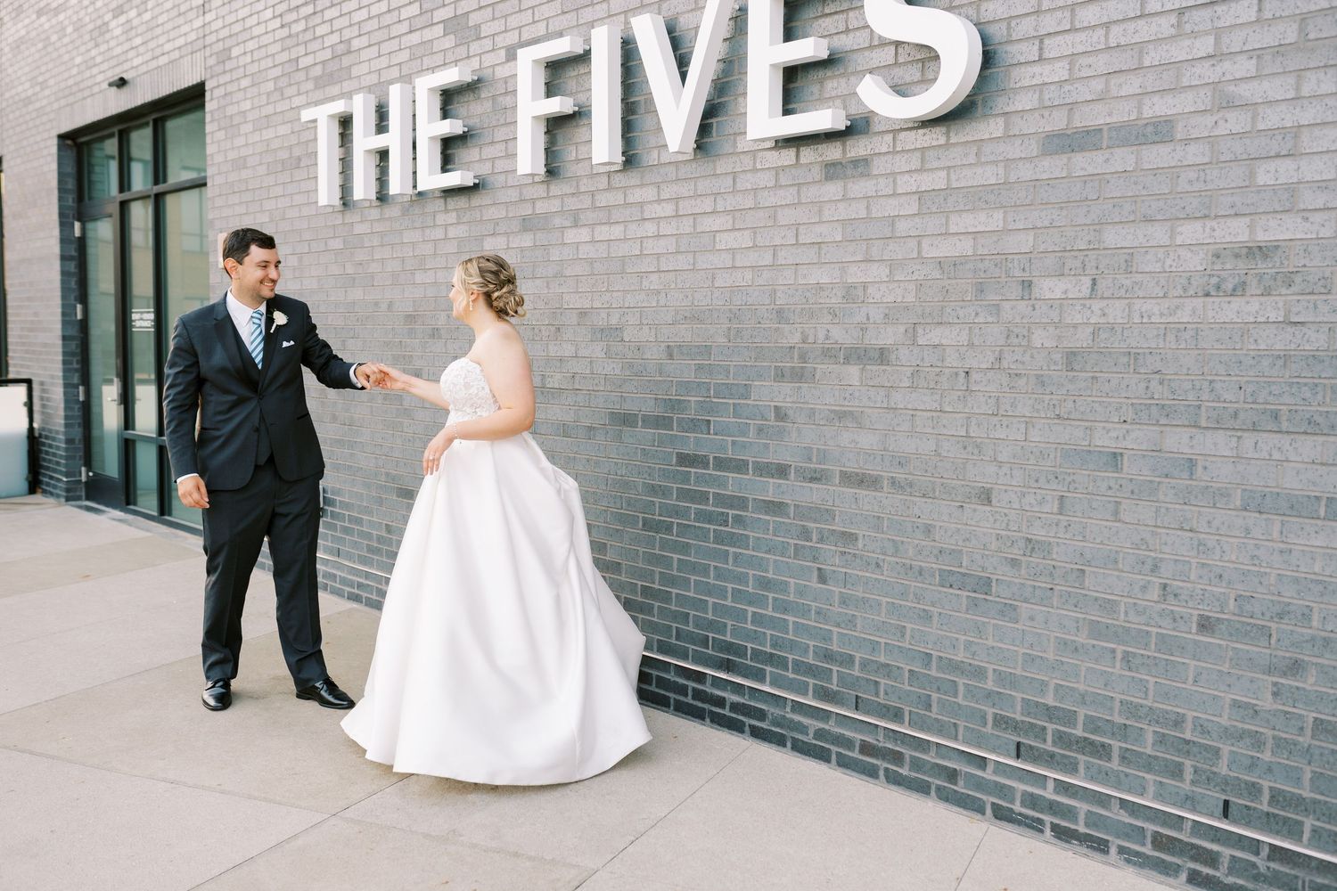 A bride and groom pose together against a gray brick wall with 'THE FIVES' text signage.