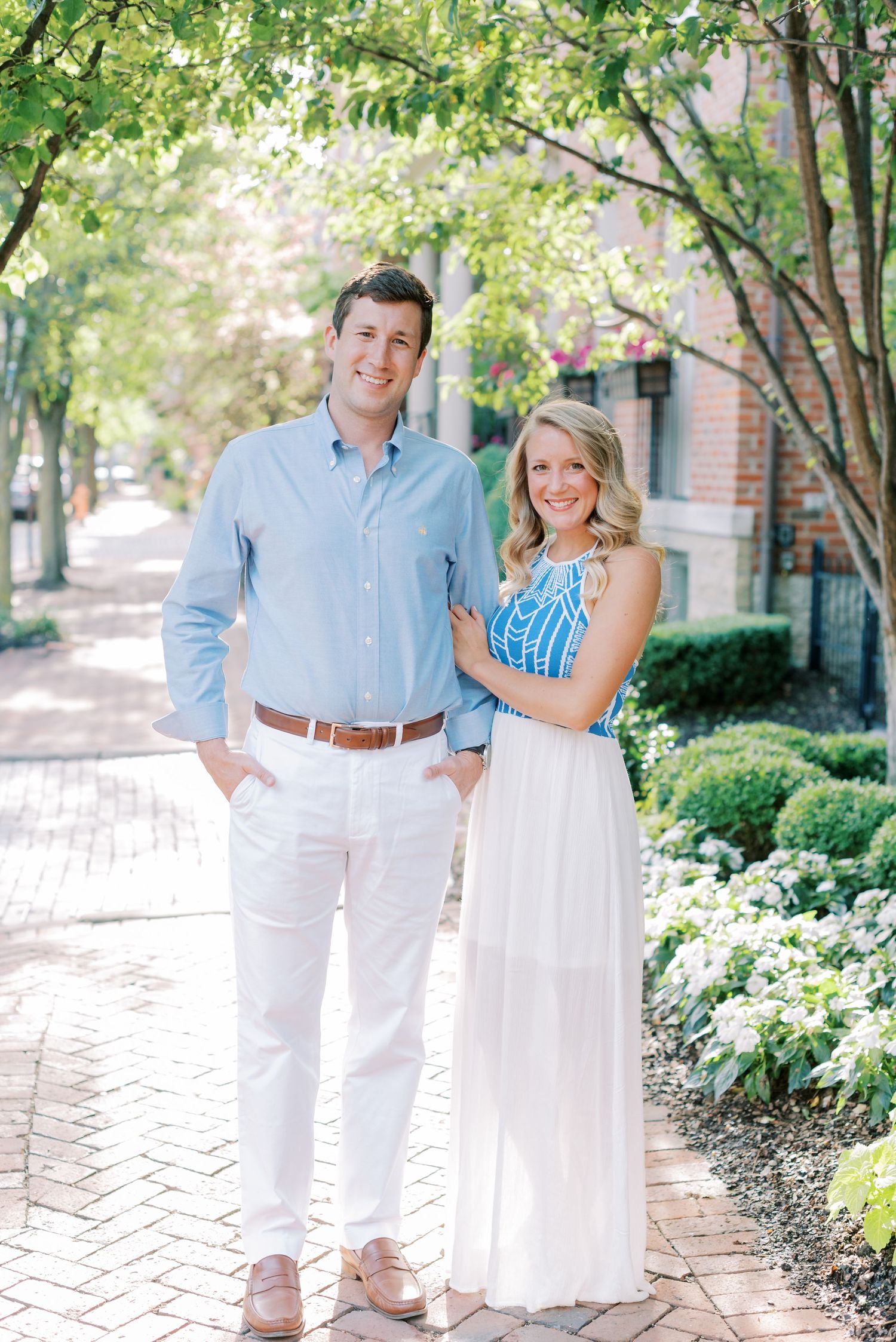 A couple in coordinating white and blue outfits poses together in a garden setting with brick pathways and greenery.