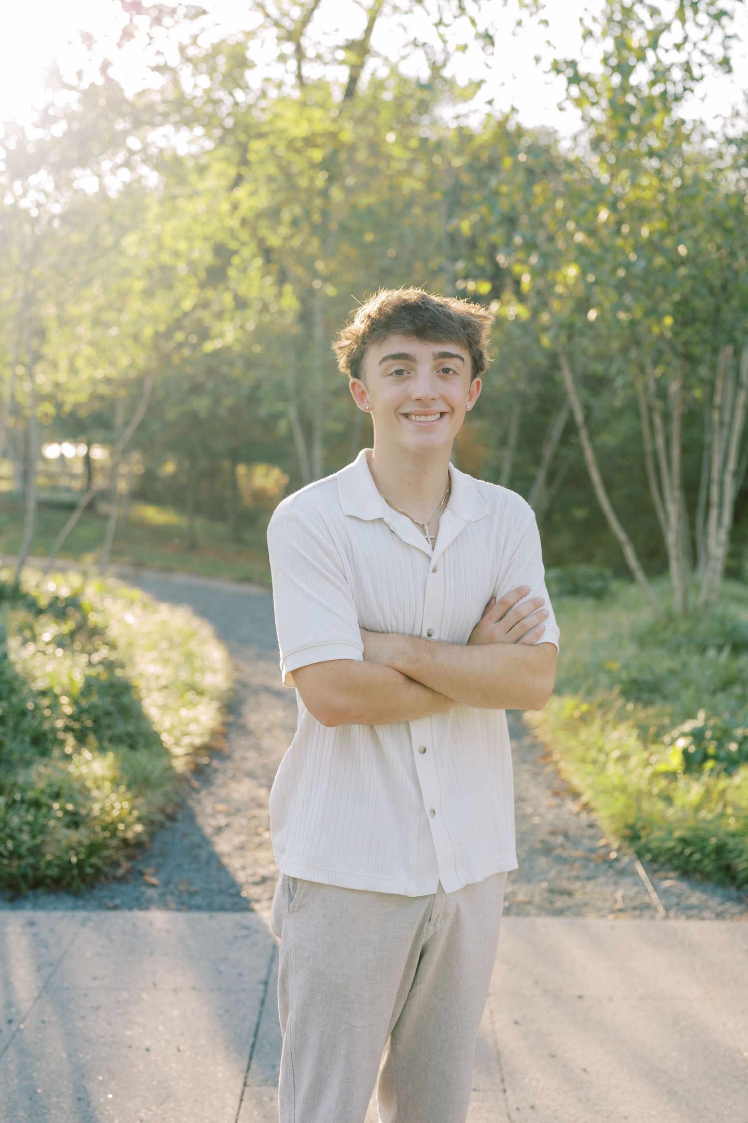 A person in a white button-down shirt and khaki pants stands on a sunlit path surrounded by greenery.
