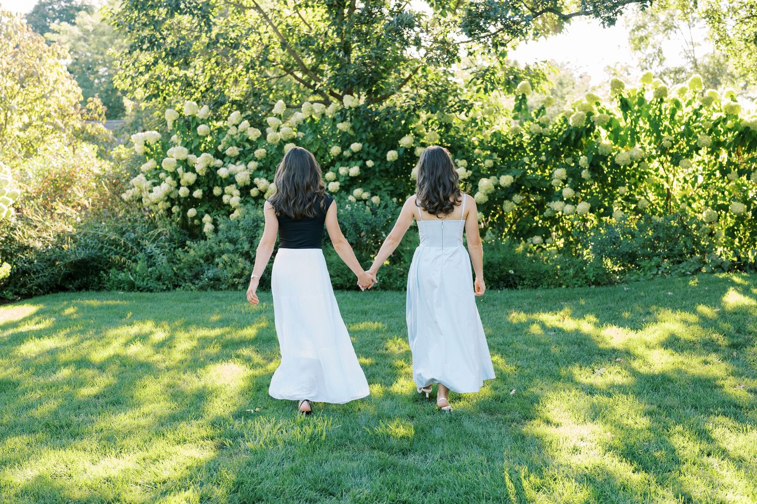 Two people in white dresses walk together through a sunlit garden with lush green trees and grass.