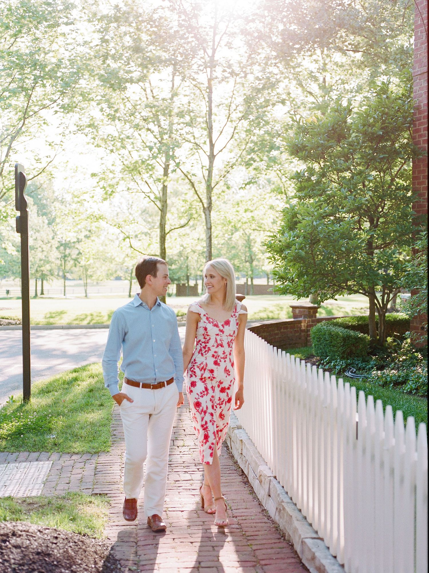 A couple strolls along a white picket fence walkway surrounded by lush greenery on a sunny summer day.
