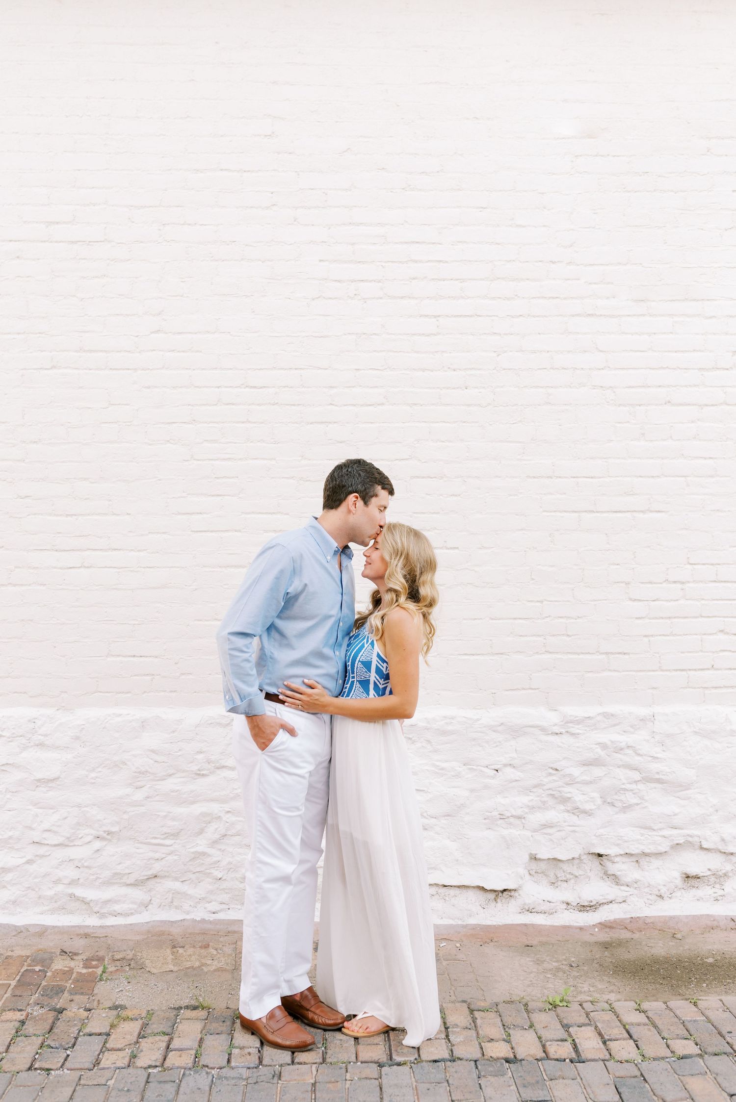 A couple shares a romantic moment on a cobblestone street beside a white wall.