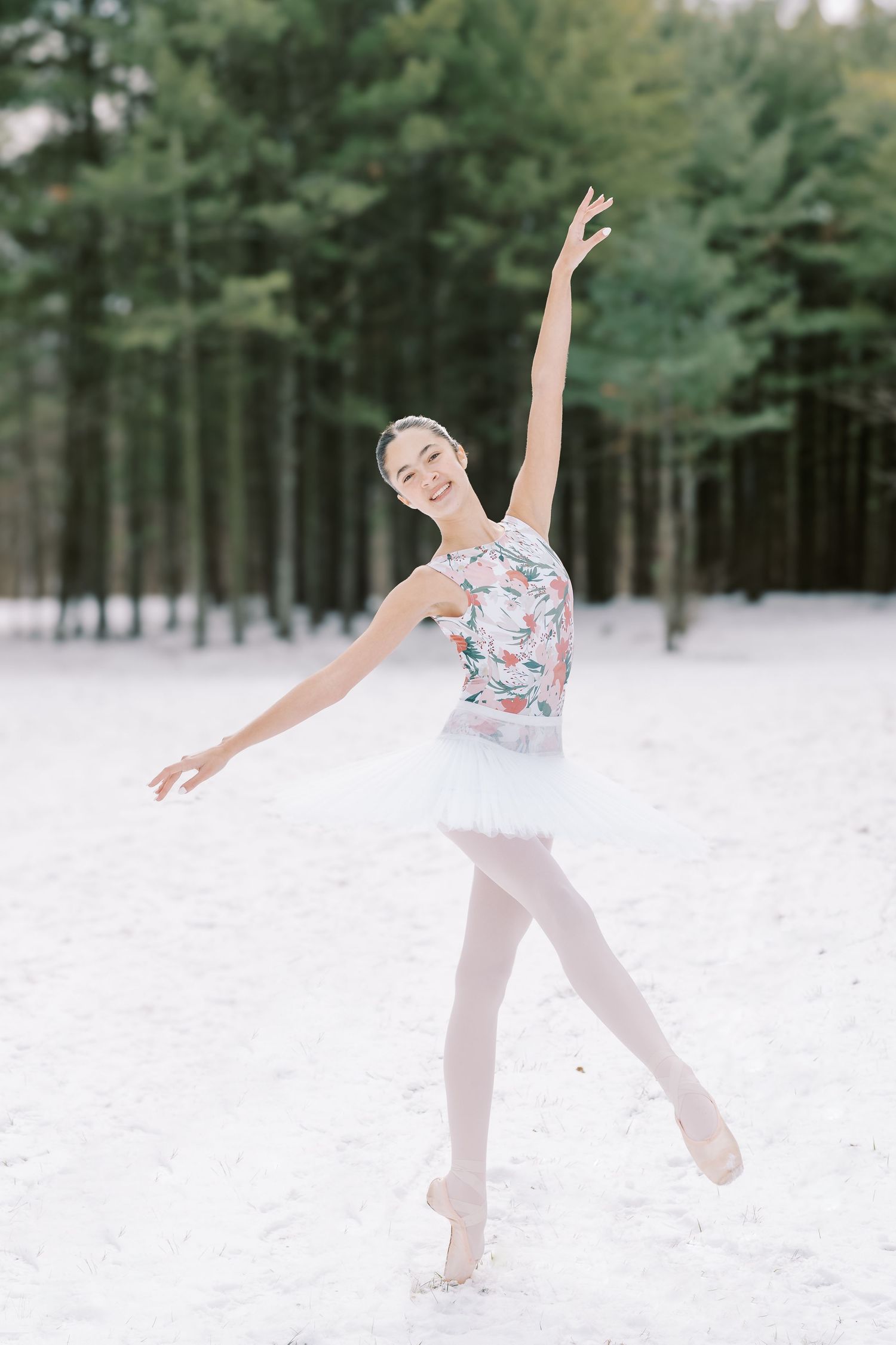 A ballet dancer in a floral leotard and white tutu performs an elegant pose on pointe in a snowy forest setting.
