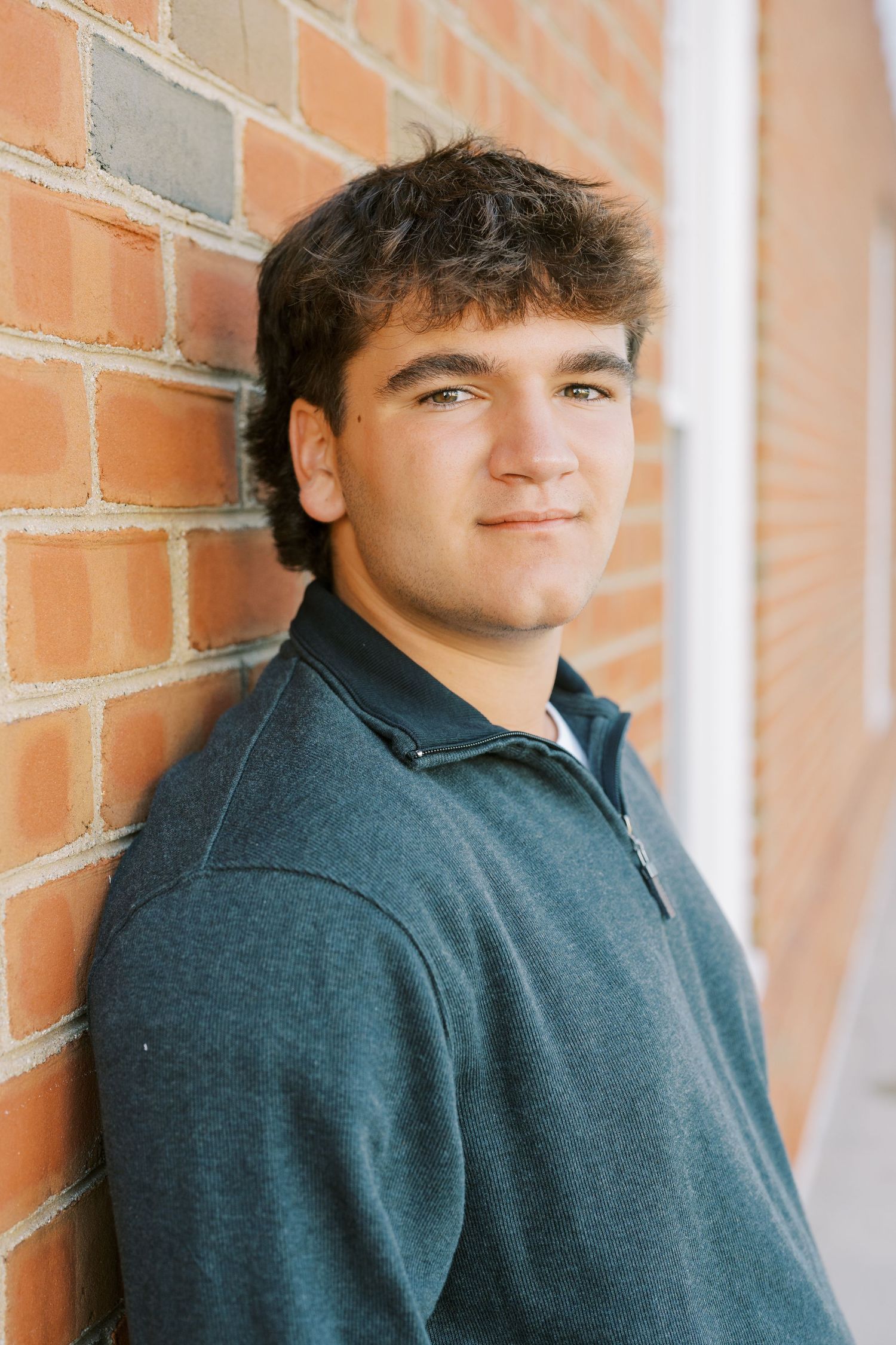 Young person in dark blue pullover smiling while leaning against a brick wall.