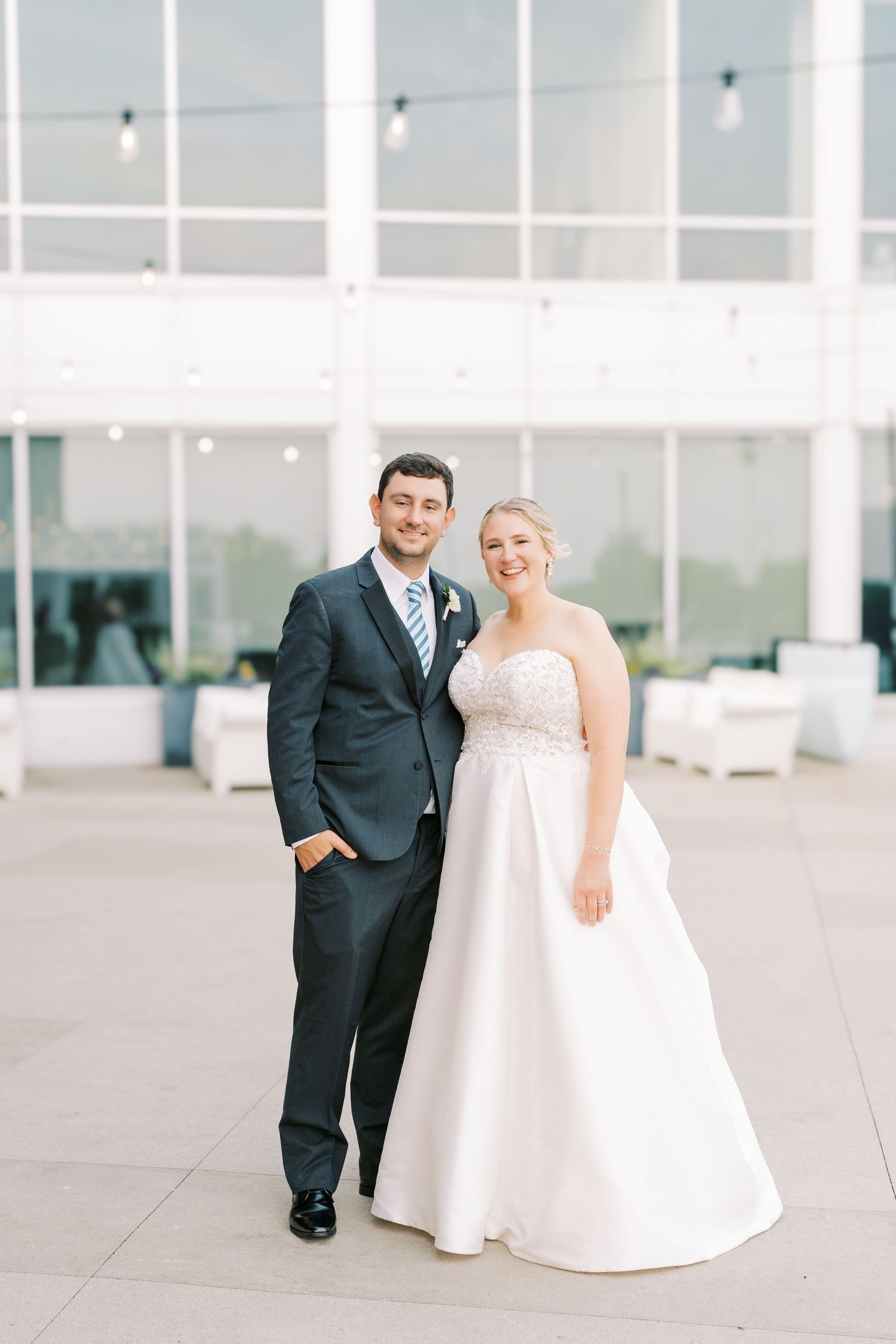 A couple poses together at their wedding in an elegant venue with white walls and string lights.