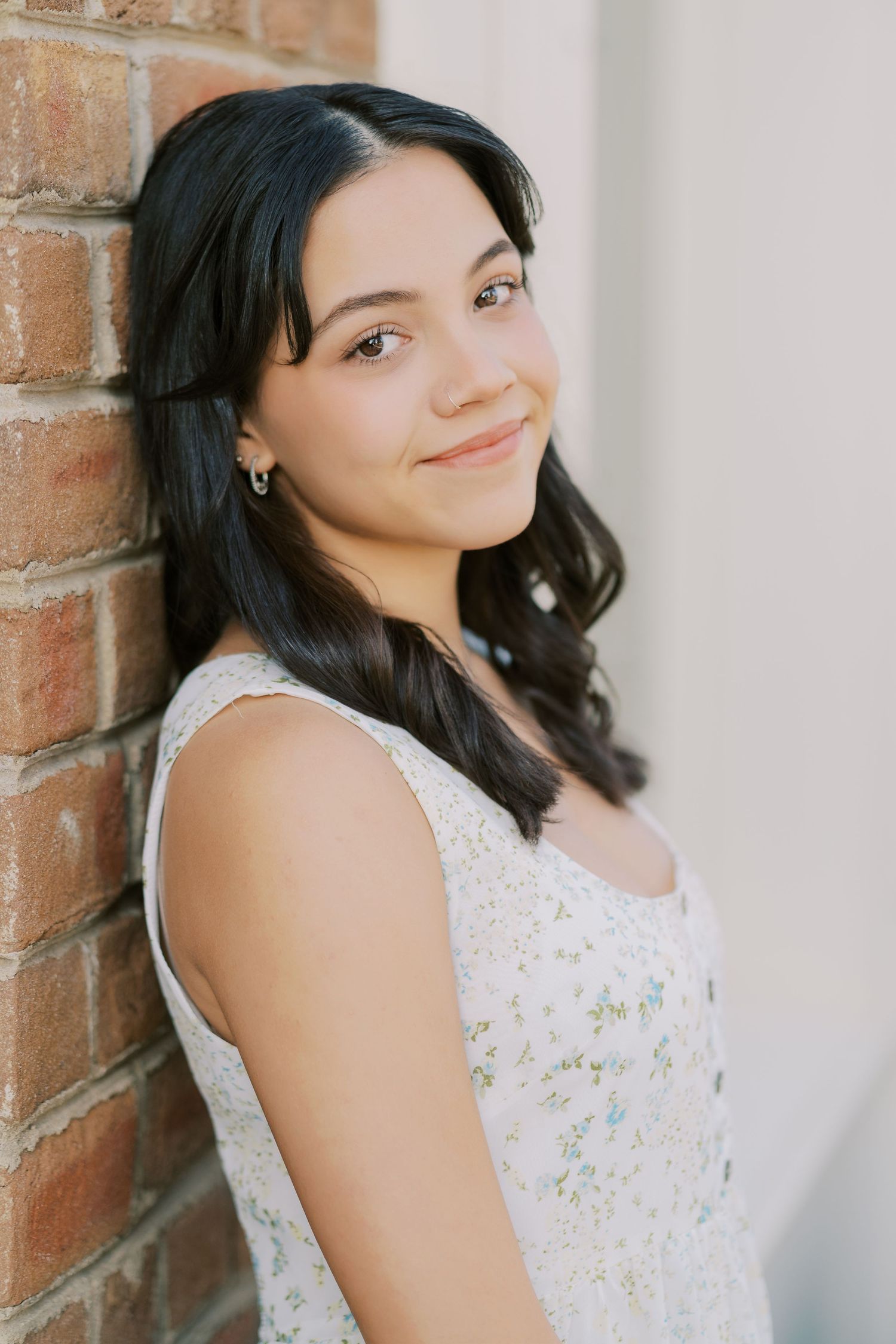 Portrait of someone in a white floral sleeveless dress leaning against a brick wall with long dark hair and a gentle smile.