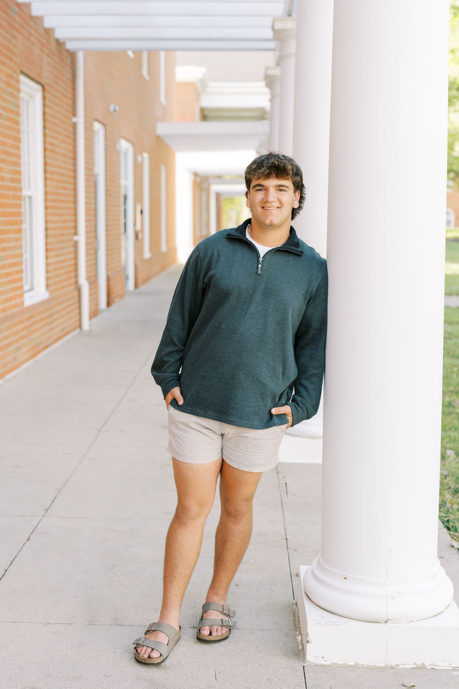 Someone wearing a dark green quarter-zip pullover and khaki shorts stands in a columned walkway on a sunny day.