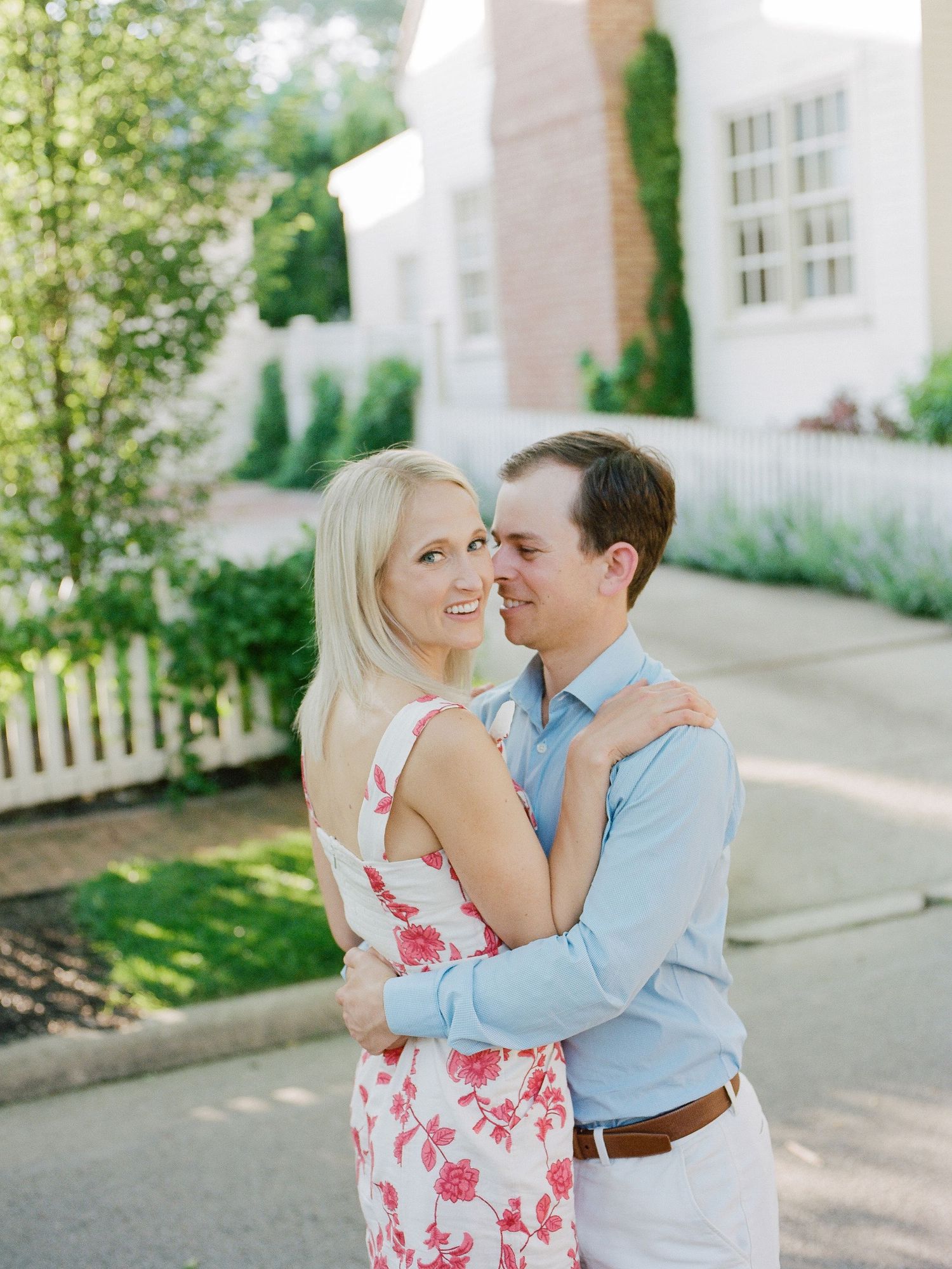 A couple embraces outdoors on a sunny day in front of a white house with a picket fence and landscaped garden.