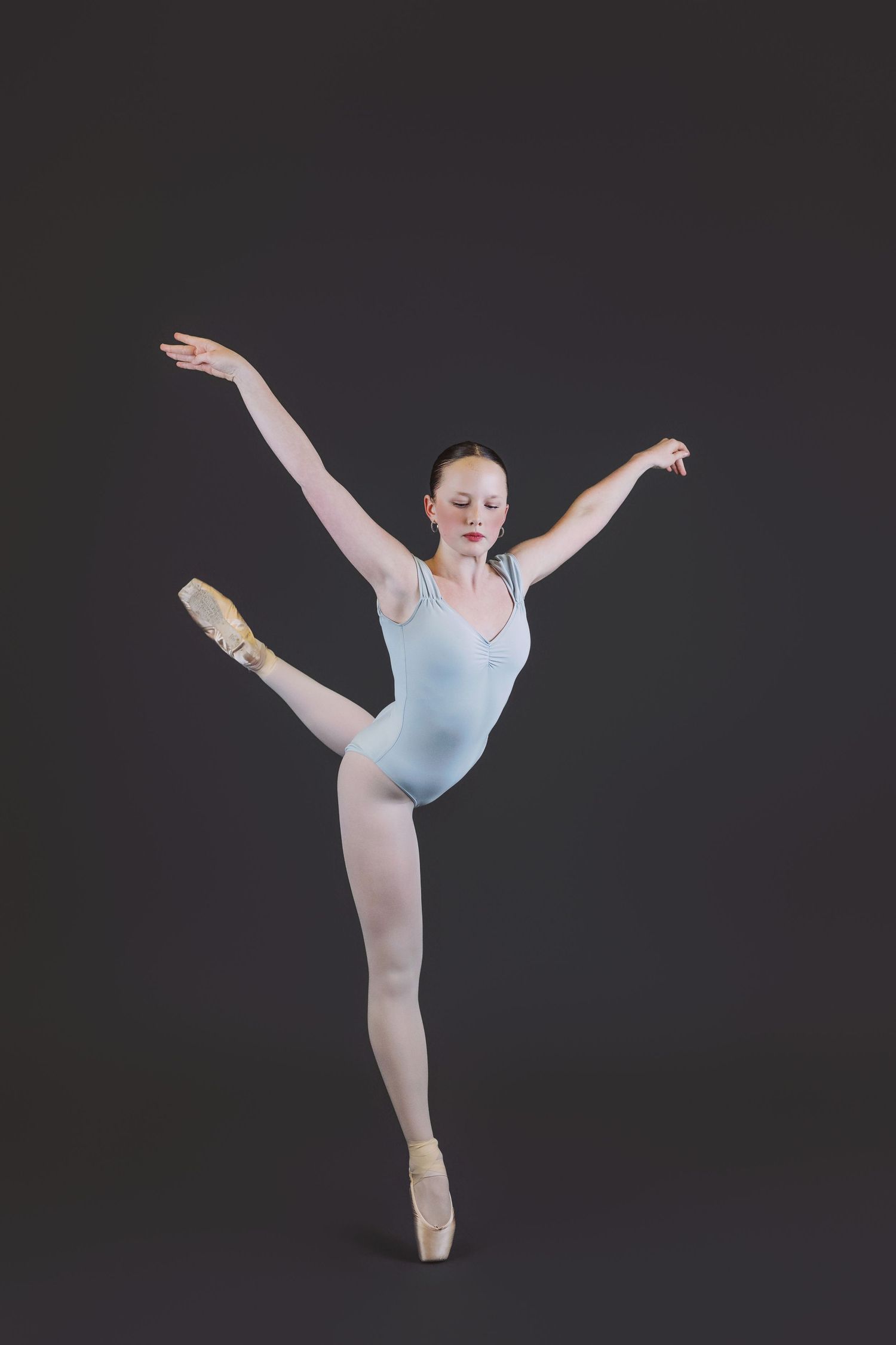 A ballet dancer in a light blue leotard performs an arabesque pose on pointe against a dark background.