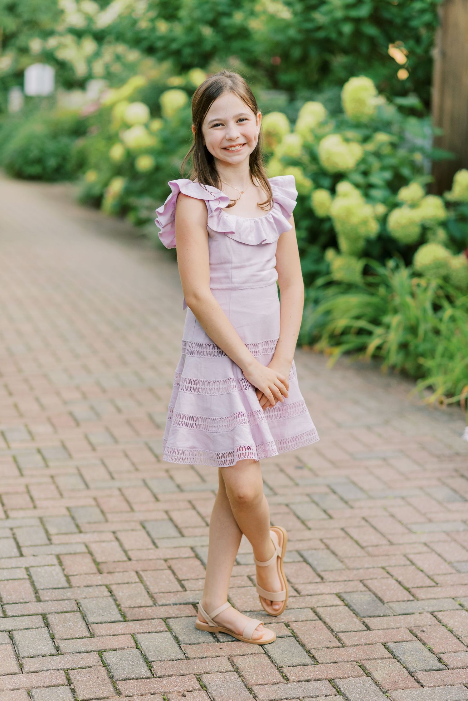 Girl in pink ruffled dress poses on brick walkway with hydrangea bushes in background.