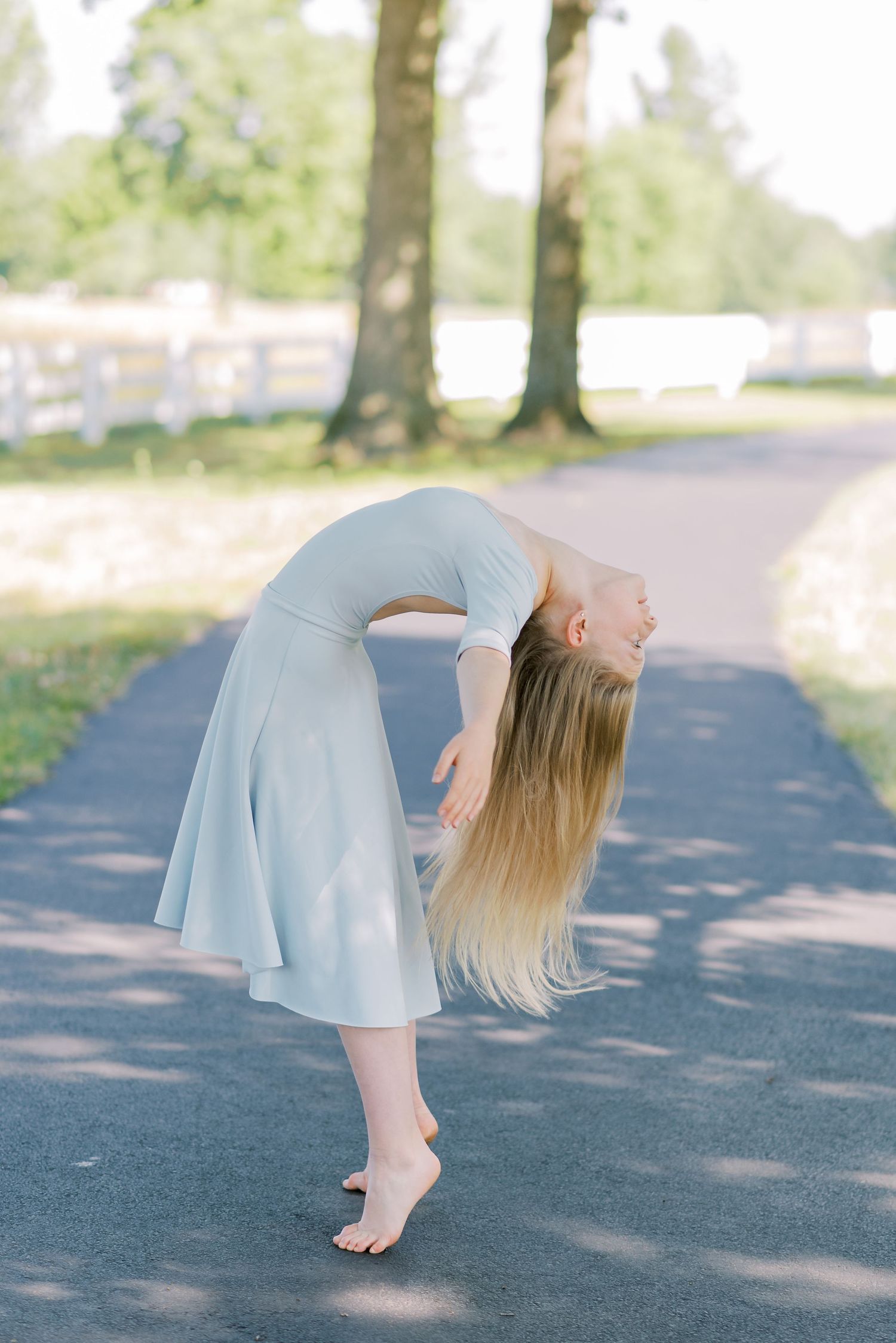 A person in a light blue dress performs a graceful dance movement on a tree-lined path.